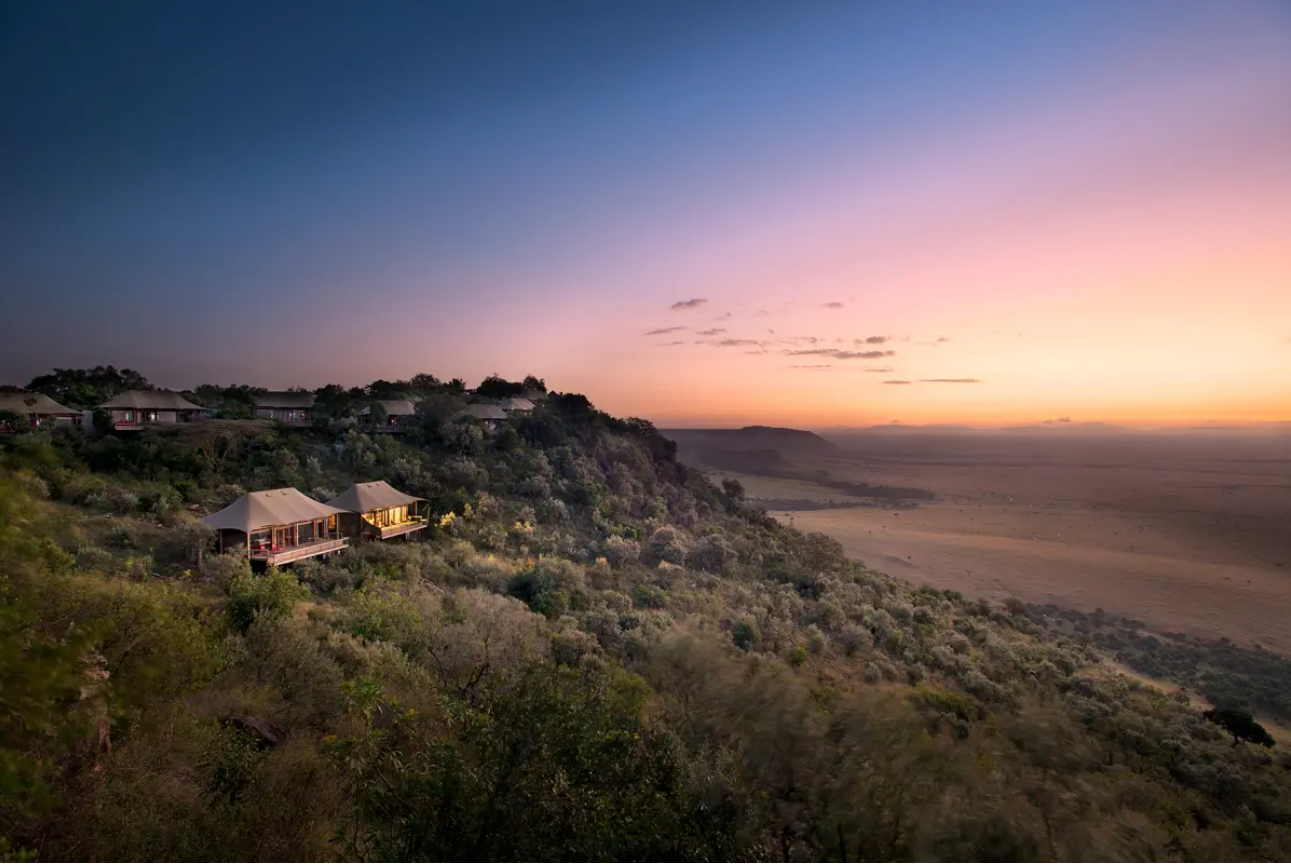 A hillside with several luxury resort buildings and tents overlooking a vast plain at sunset, with pink and purple sky.