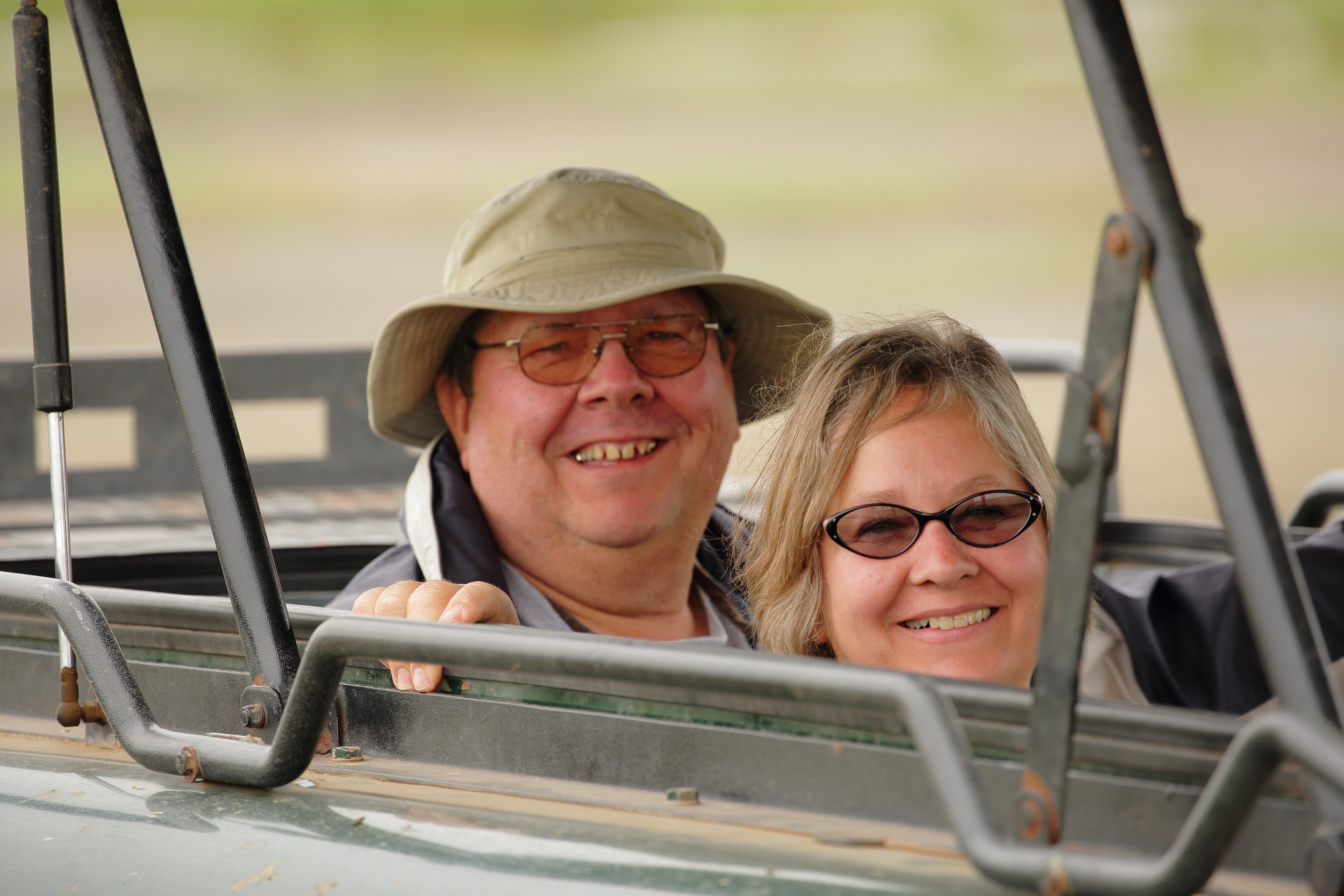 A smiling man and woman wearing sunglasses sitting in a vehicle outdoors.