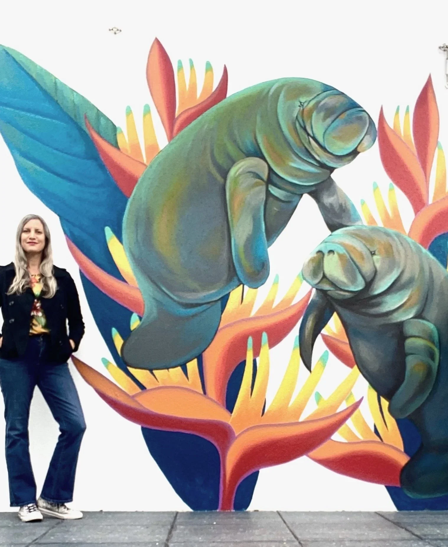 Woman standing in front of vibrant mural of two talking frogs with large blue and green leaves and red and orange flowers.