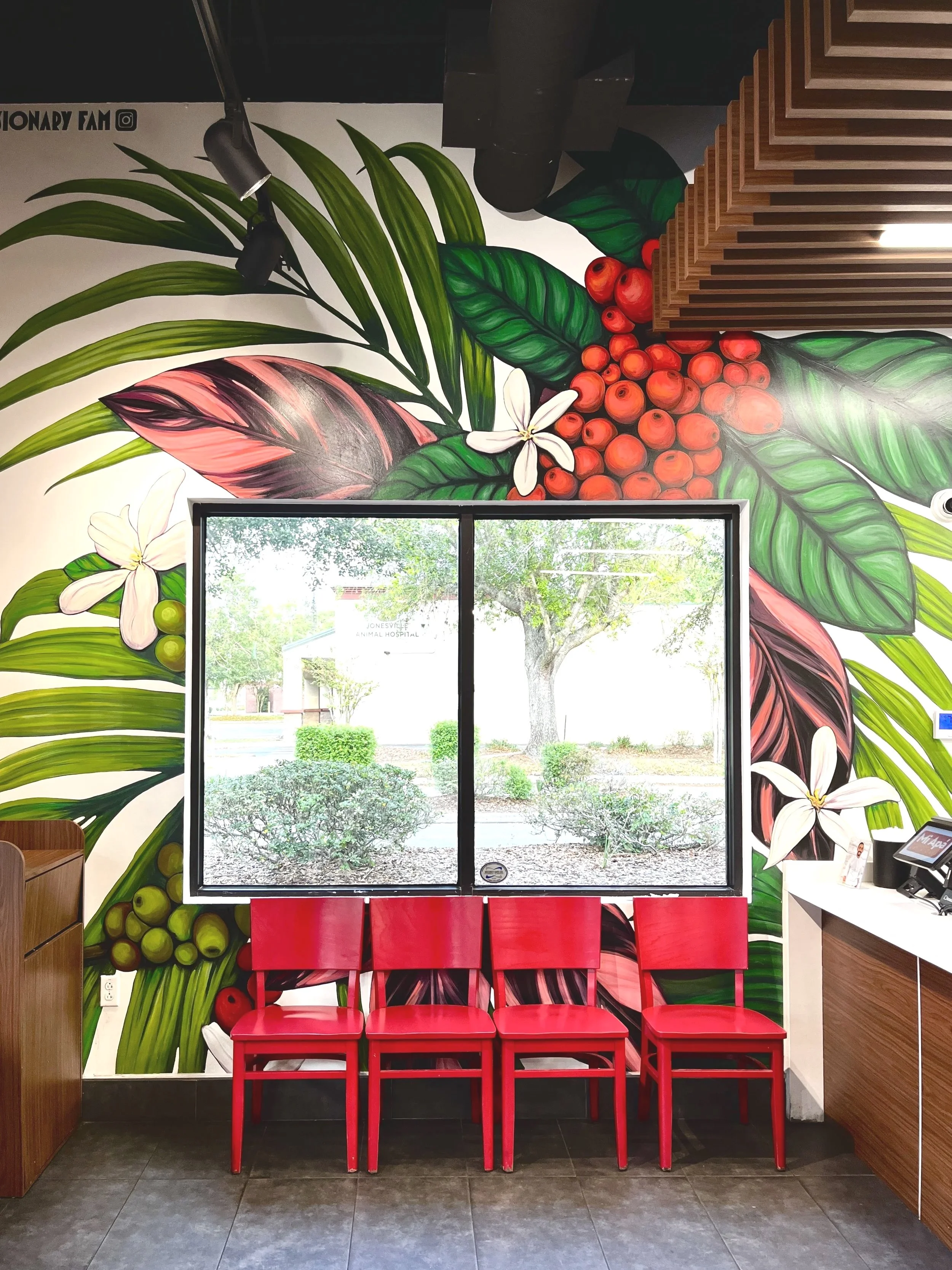 Colorful mural of tropical leaves, red berries, and white flowers on a wall behind four red chairs in a waiting area near a window showing trees and bushes outside.