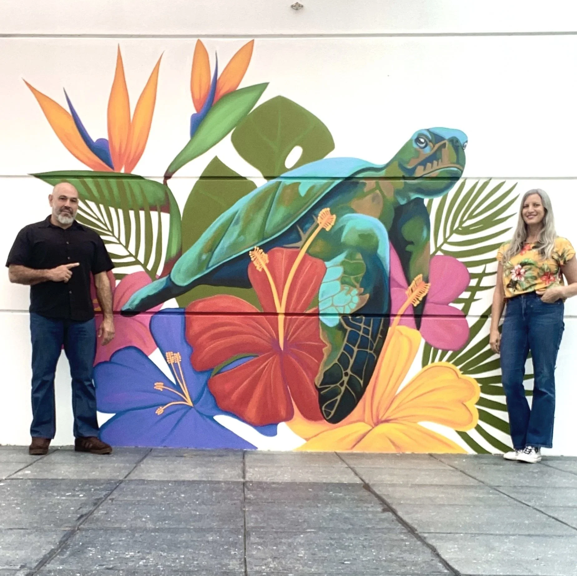 A mural featuring colorful tropical flowers and a green chameleon with a background of green leaves, with two people standing on either side of it.