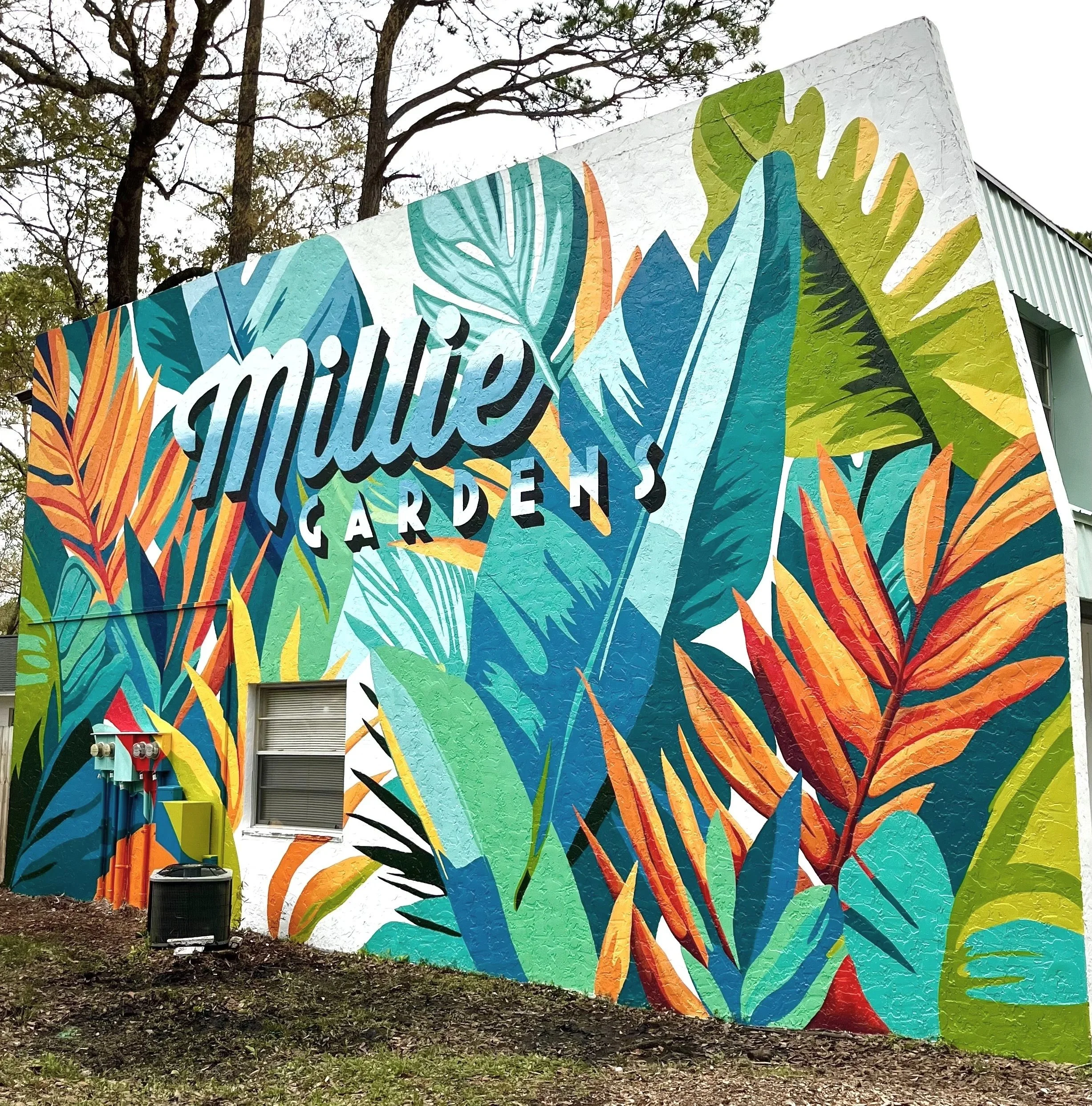 Colorful mural on a building exterior displaying the words 'Mullie Gardens' surrounded by vibrant tropical leaves and plants.