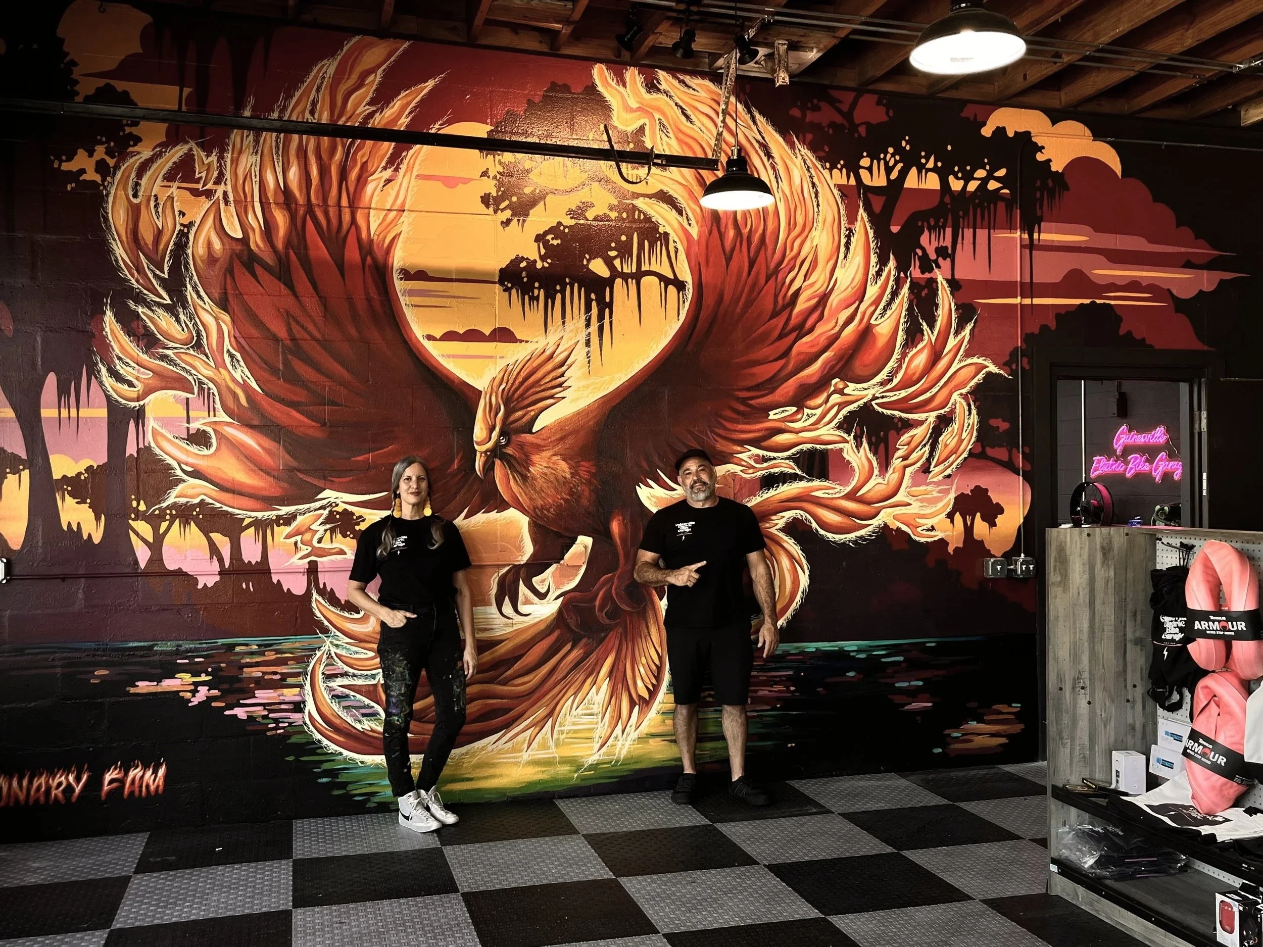 Two people standing in front of a vibrant mural of a phoenix with wings spread, rising above a body of water with trees and sunset in the background.