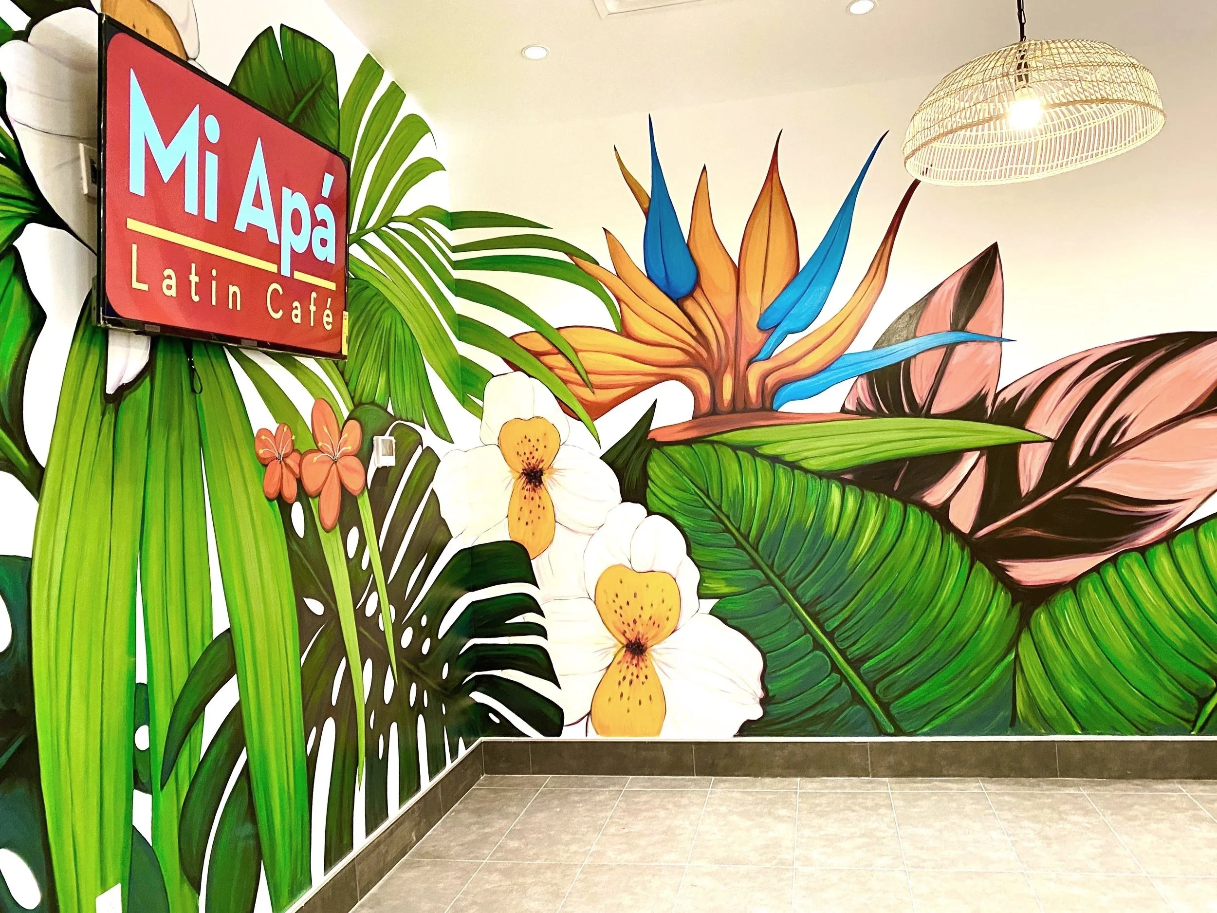 Colorful mural of tropical plants and flowers inside a cafe with a red sign that says 'Mi Apá Latin Café' in the corner.