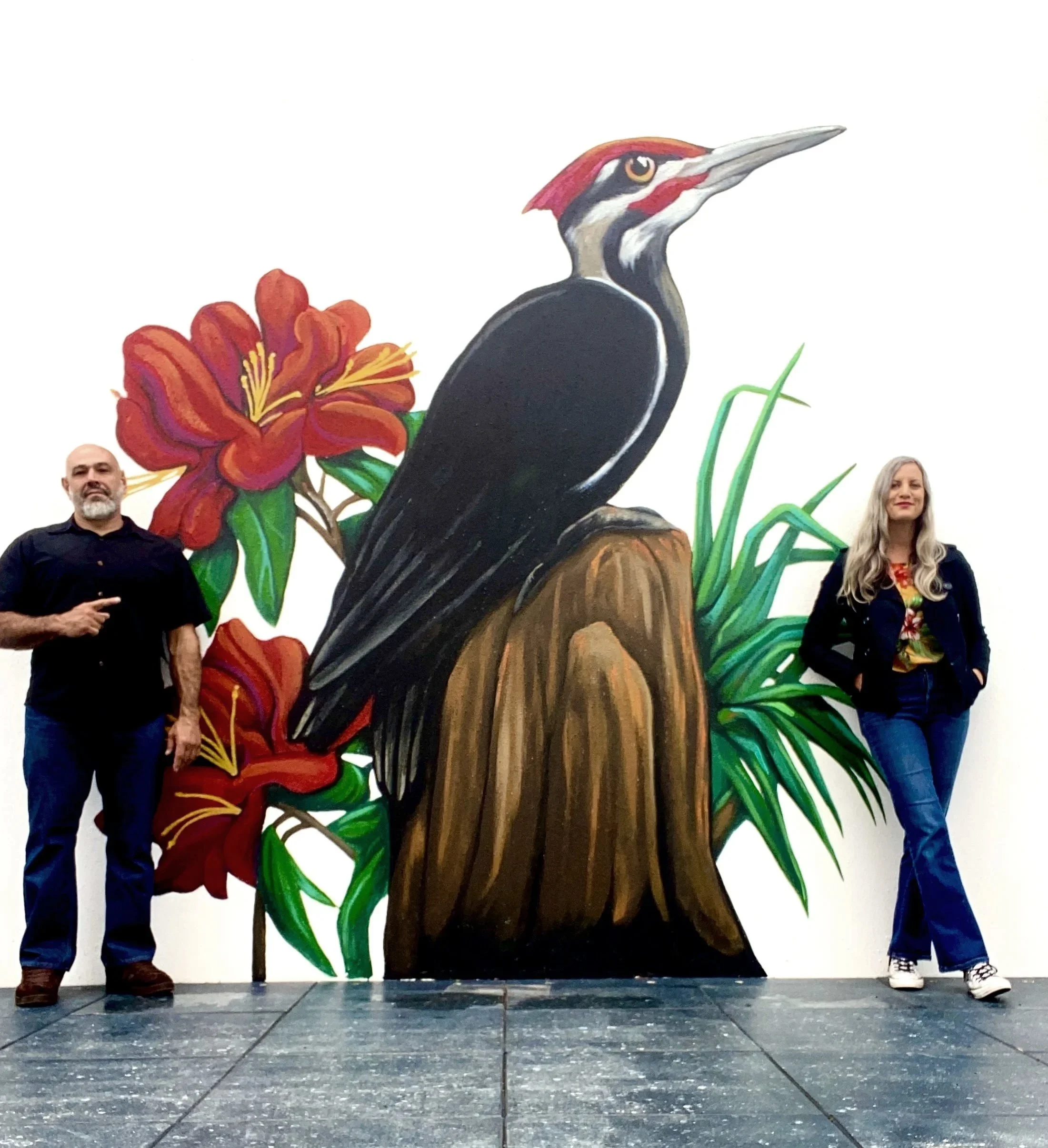 Man and woman standing on either side of a large mural of a woodpecker with red flowers and green foliage.