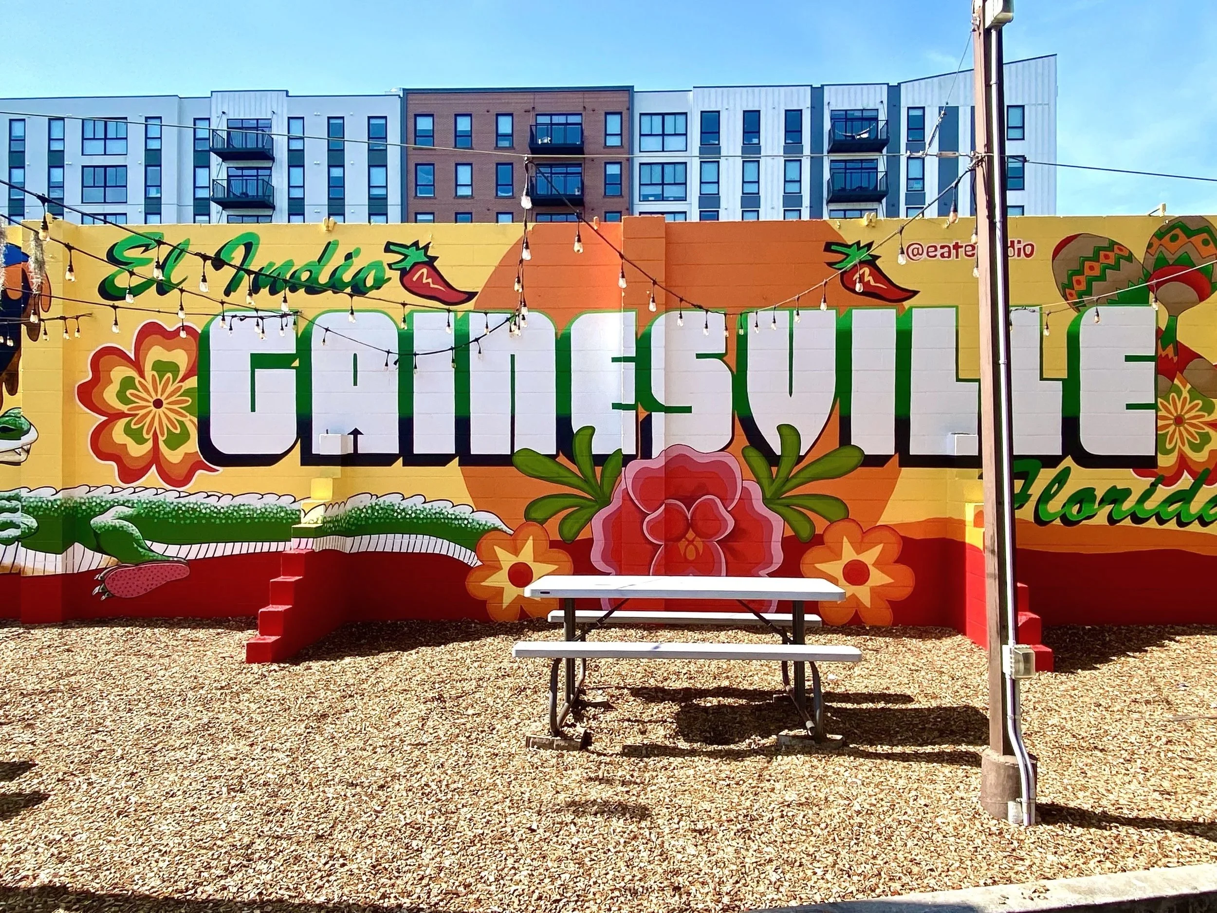 Colorful mural reading 'El Audio' and 'Cali Sugar Florida' on a yellow wall, decorated with flowers and chili peppers, with string lights hanging above. In front, there is a white picnic table on gravel ground.