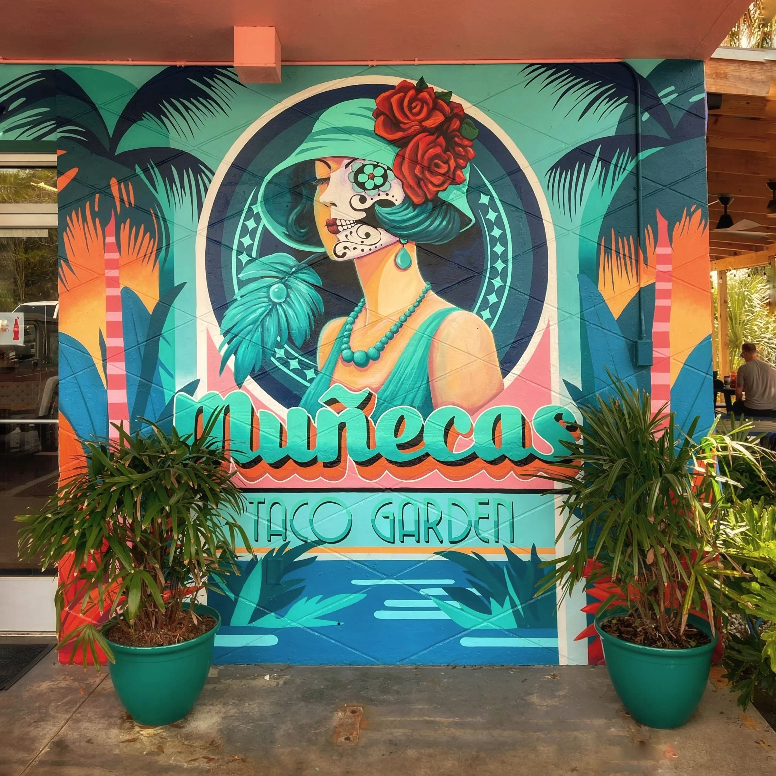 Colorful mural featuring a woman with teal hair, floral headpiece, and sugar skull face makeup, with the text 'Muñecas Taco Garden' at the bottom, surrounded by tropical plants.