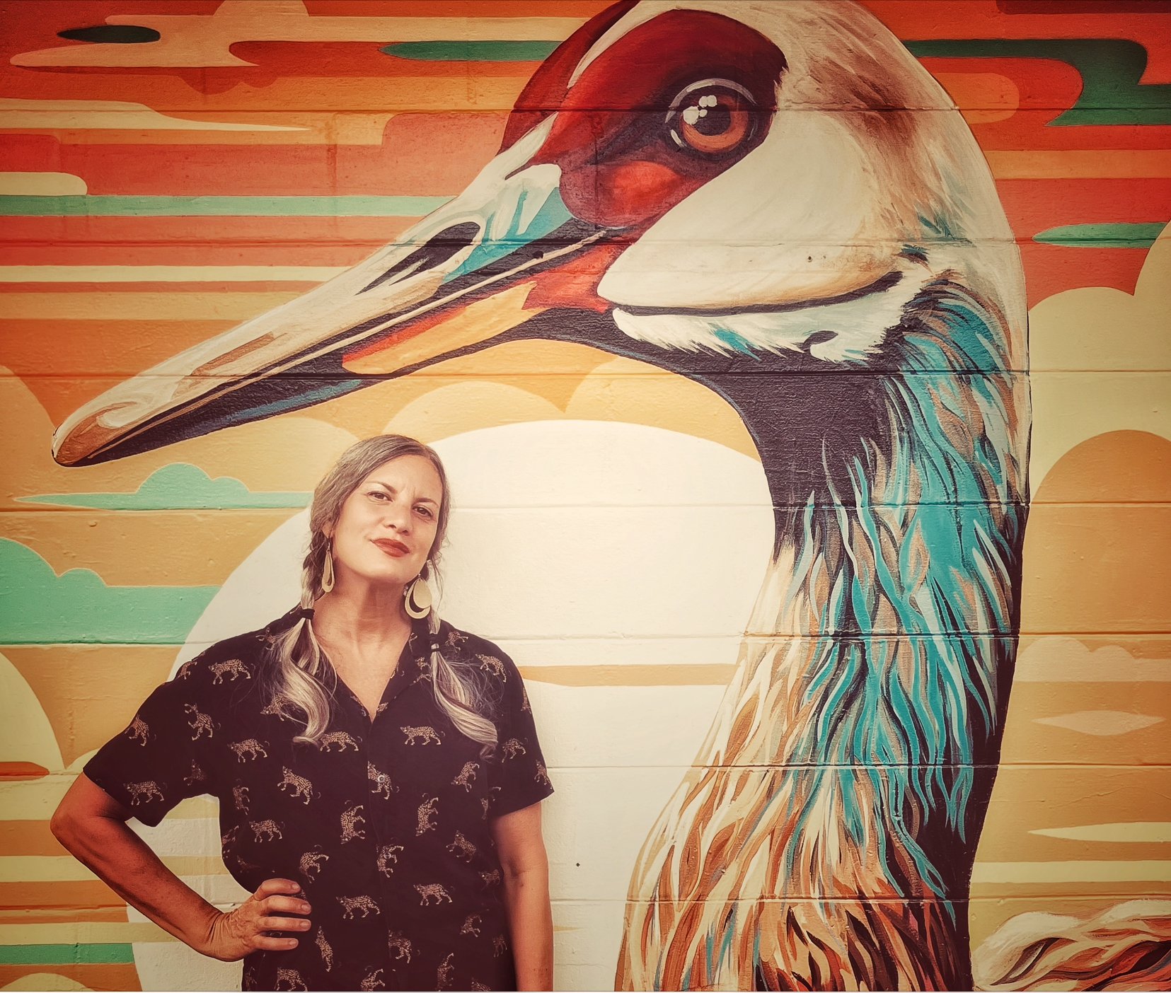 A woman with long, gray hair styled in pigtails, wearing a black shirt with a tiger pattern, standing in front of a colorful mural of a large bird's head, possibly a heron or crane, with a fiery orange and yellow background.