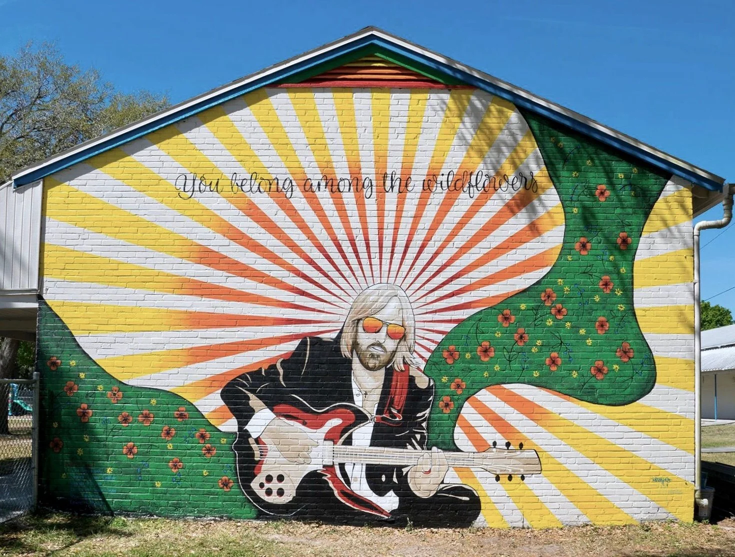 Mural of a man with long blond hair and sunglasses playing an electric guitar, with a yellow and orange radiating sunburst background and green foliage with red flowers, and the text "You belong among the wildflowers" above.