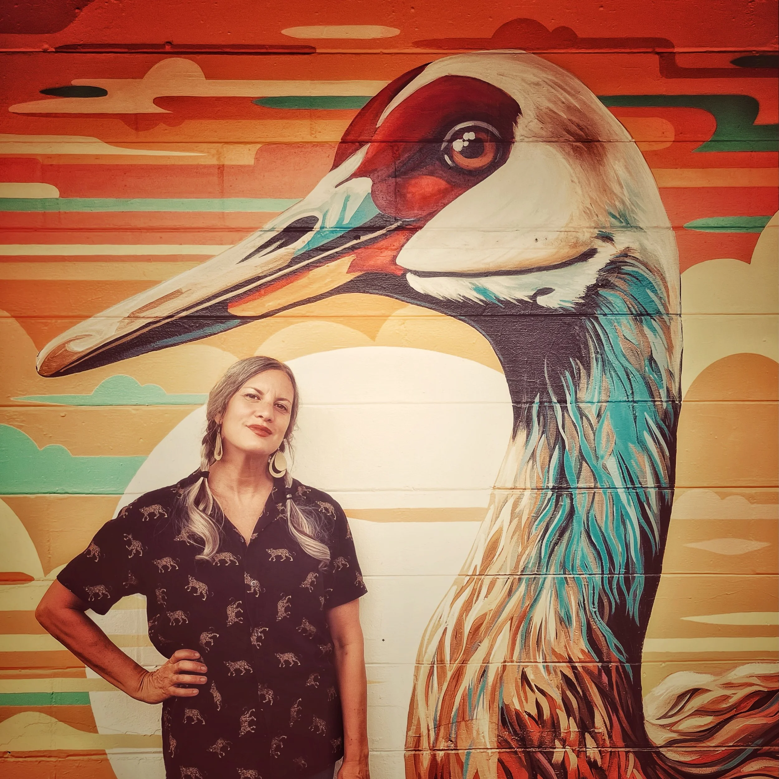 A woman with long, wavy hair wearing a black shirt with a leopard print stands in front of a large, colorful mural of a pelican against a sunset background