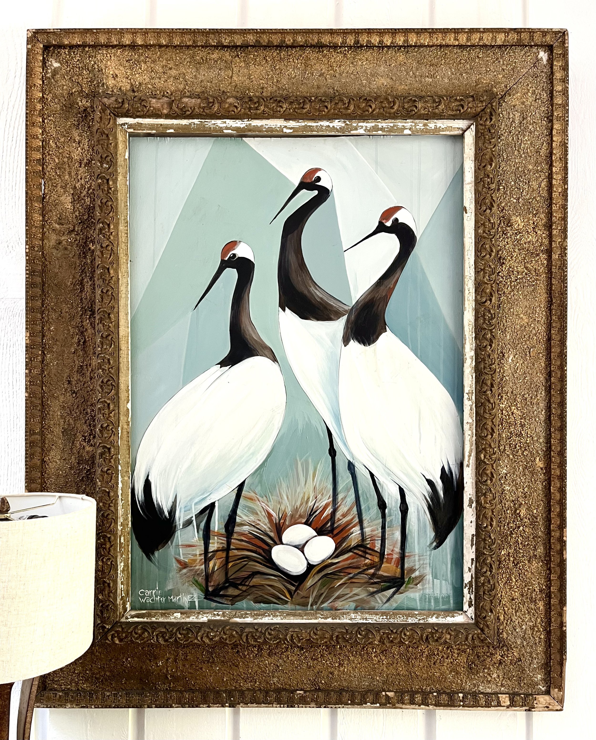 A painting of three cranes with white bodies, black wings, long black beaks, and red patches on their heads, standing in a nest with three white eggs, framed with an ornate gold frame.