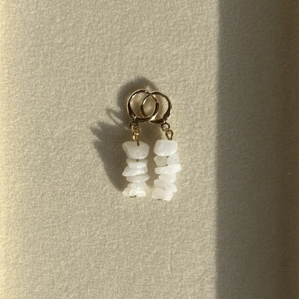 A pair of earrings with small white stones hanging from gold hoops, displayed against a beige background.
