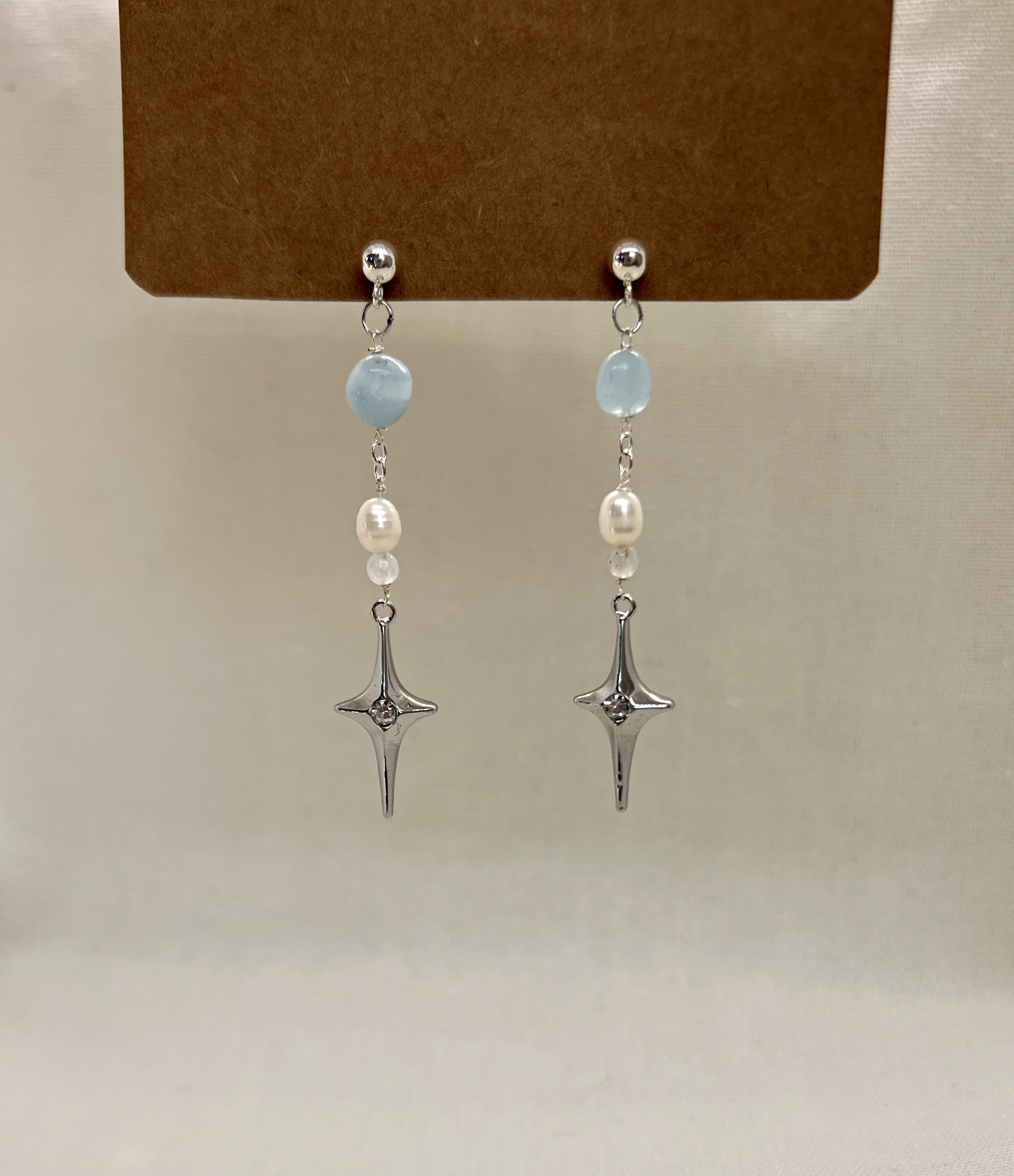 A pair of dangling earrings with light blue beads, pearls, and silver star-shaped charms, displayed on a brown card.