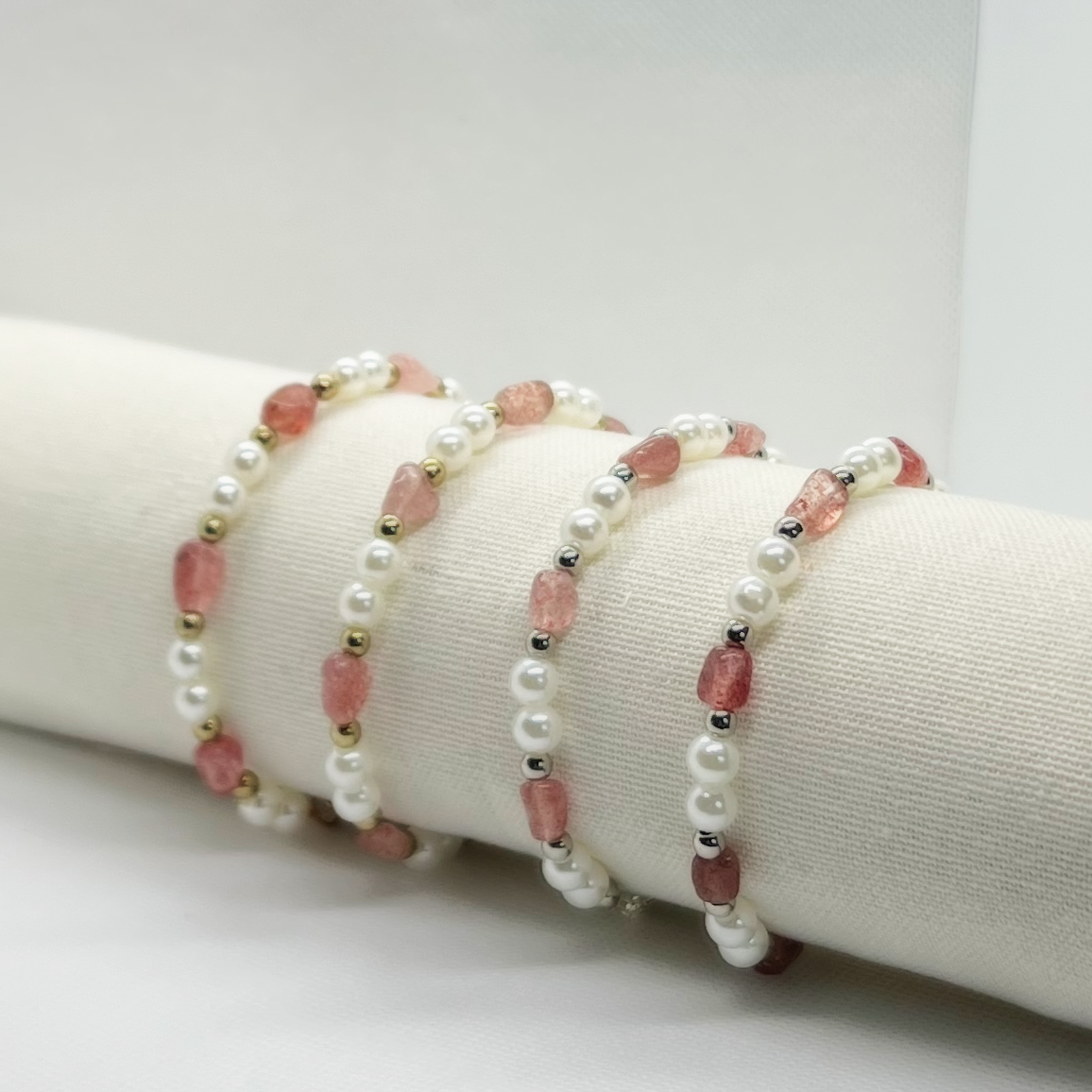 Three pearl and pink gemstone bead bracelets with metallic accents, displayed on a white cushioned surface.