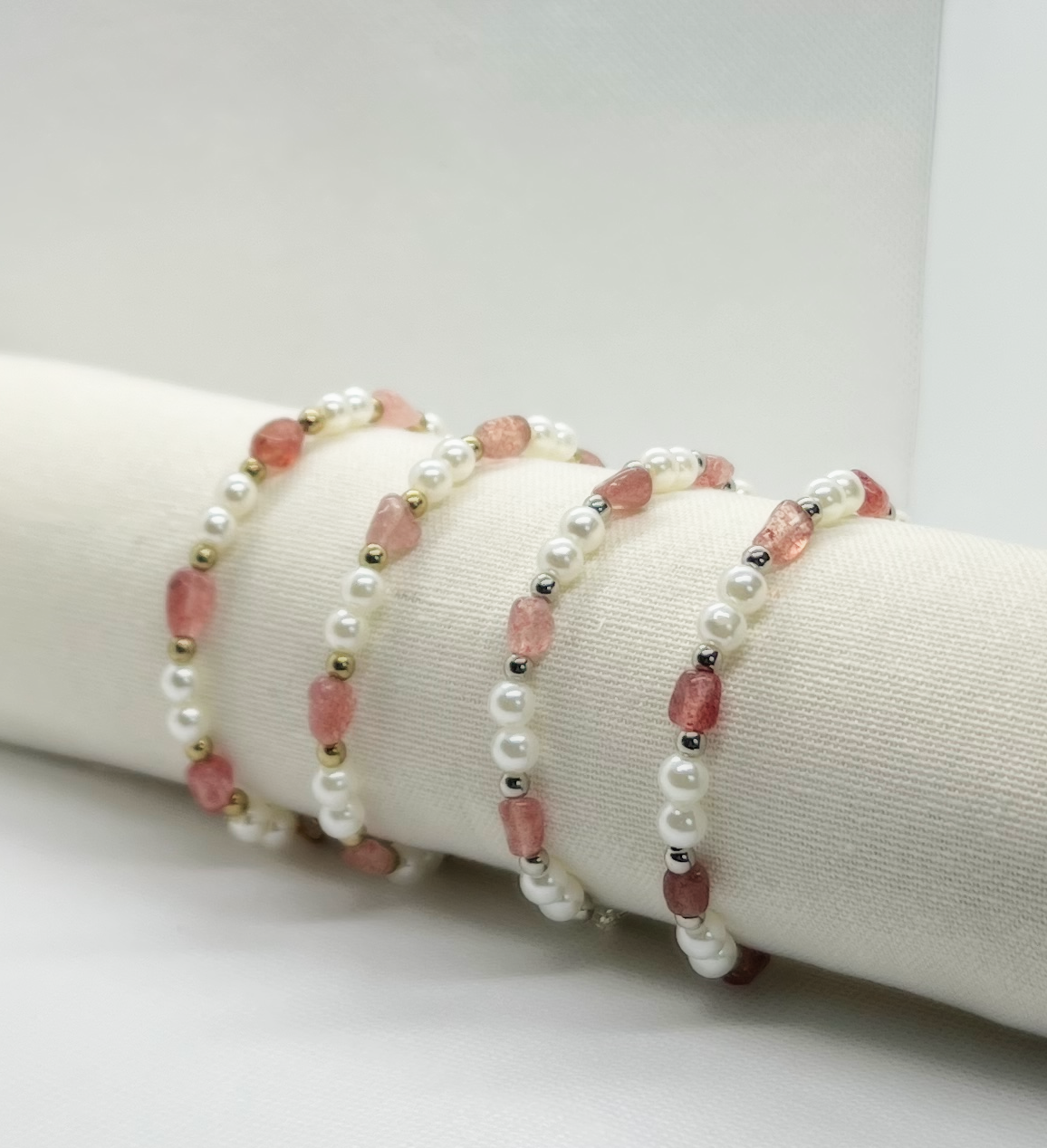 Four pearl and pink gemstone beaded bracelets displayed on a white cylindrical stand.