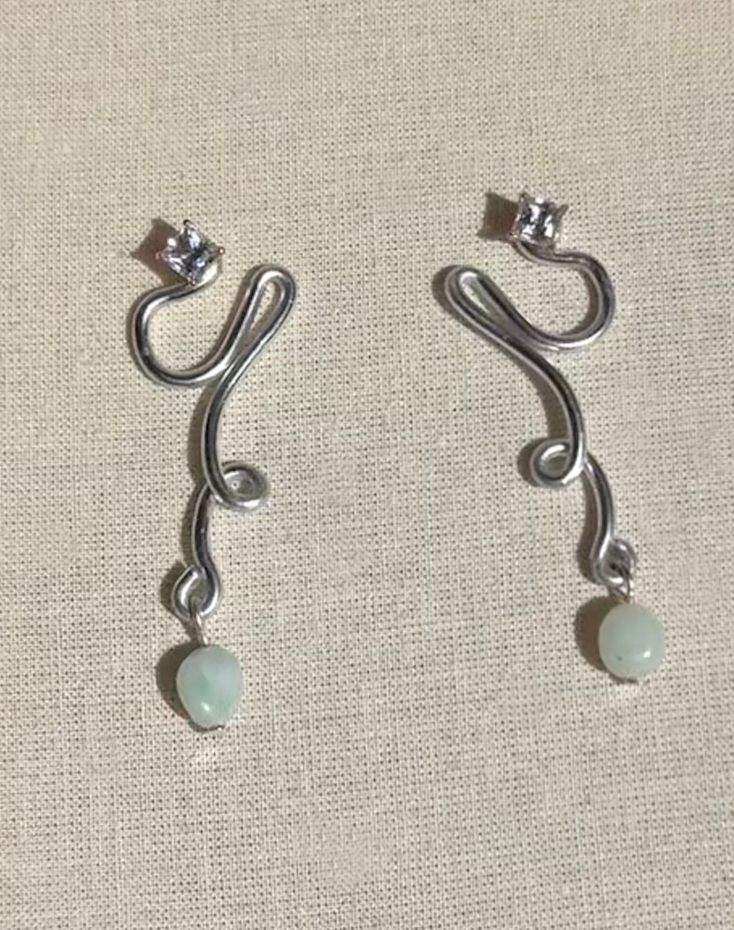 Silver earrings with a curved, wavy design and semi-precious stone dangling at the bottom, placed on a beige fabric surface.