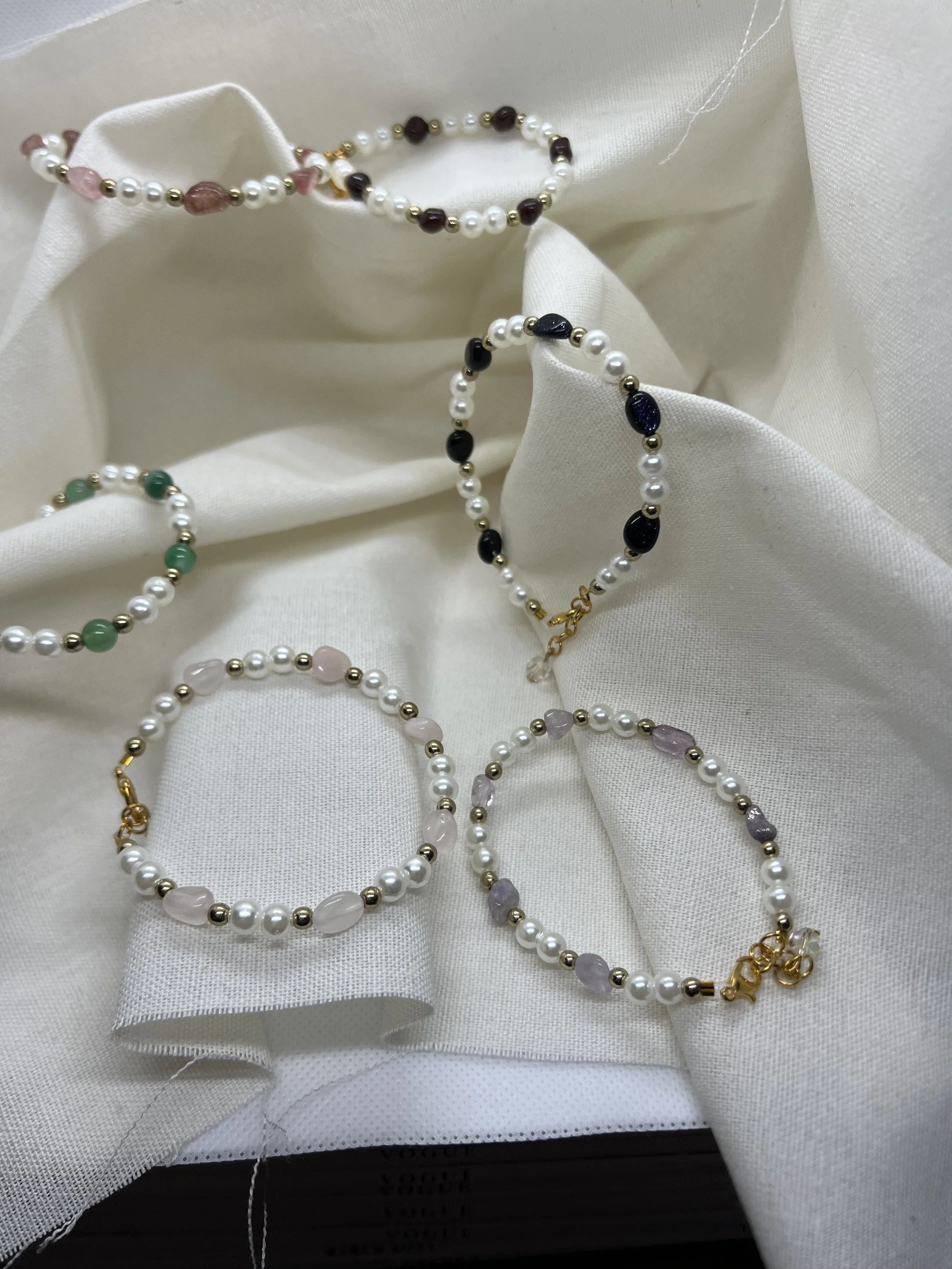 Collection of pearl and beaded jewelry on white fabric, including bracelets and necklaces with various colored beads and gold accents.