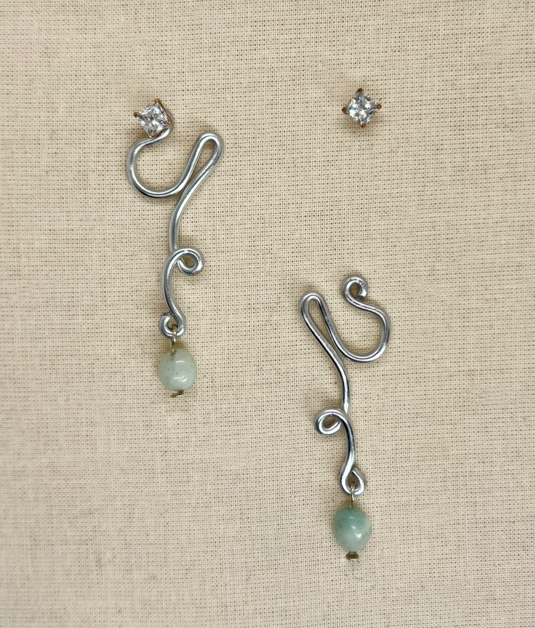 Silver dangling earrings with a light green bead at the bottom and a small clear diamond at the top, set on beige fabric.