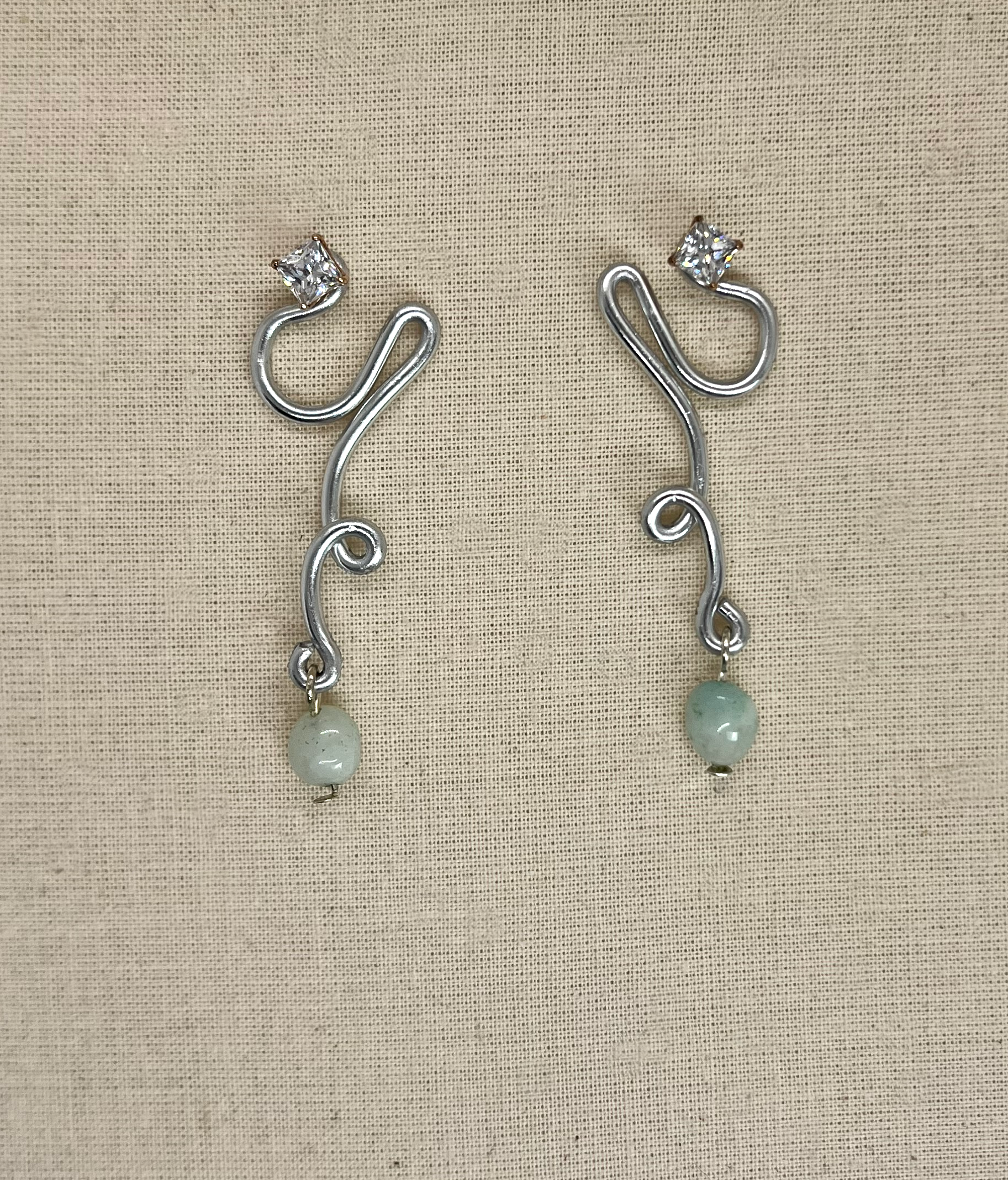 Silver earrings with a swirling design, each featuring a square gemstone at the top and a rounded, pale green bead hanging at the bottom.