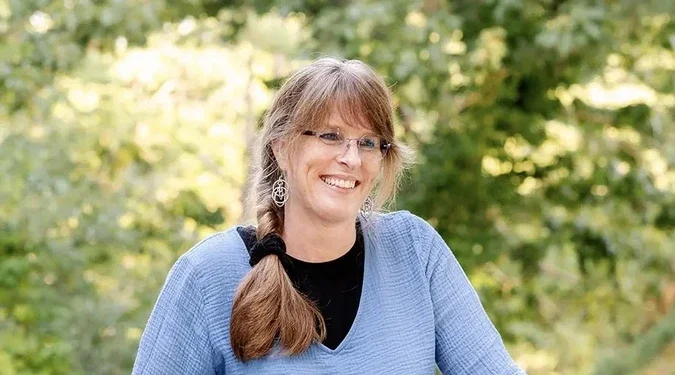 A woman with glasses and a braid smiling outdoors, wearing a blue sweater and earrings, with greenery in the background.