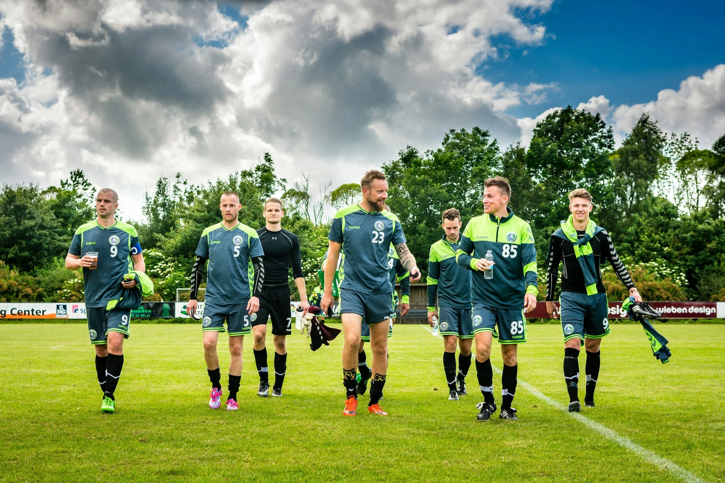 A group of seven male soccer players in navy and neon green uniforms walking on a soccer field, carrying drinks and gear, with a backdrop of trees and a partly cloudy sky.