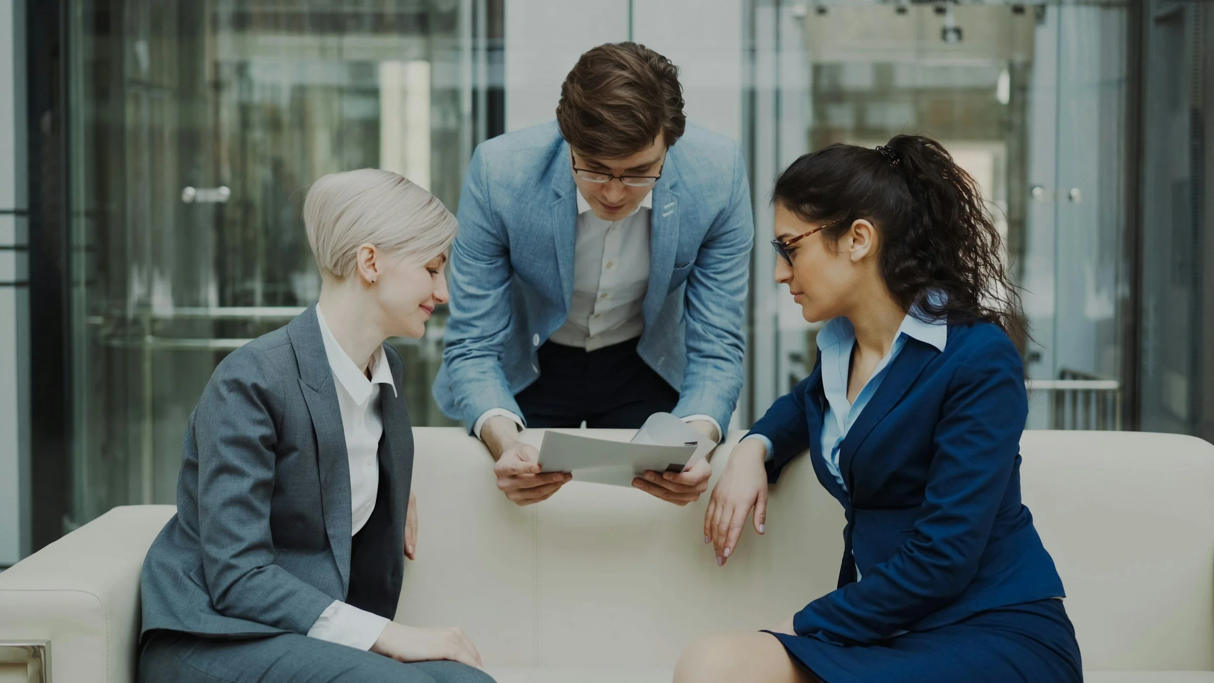Two women in business suits sitting on a couch and a man standing, all looking at documents together in an office setting.