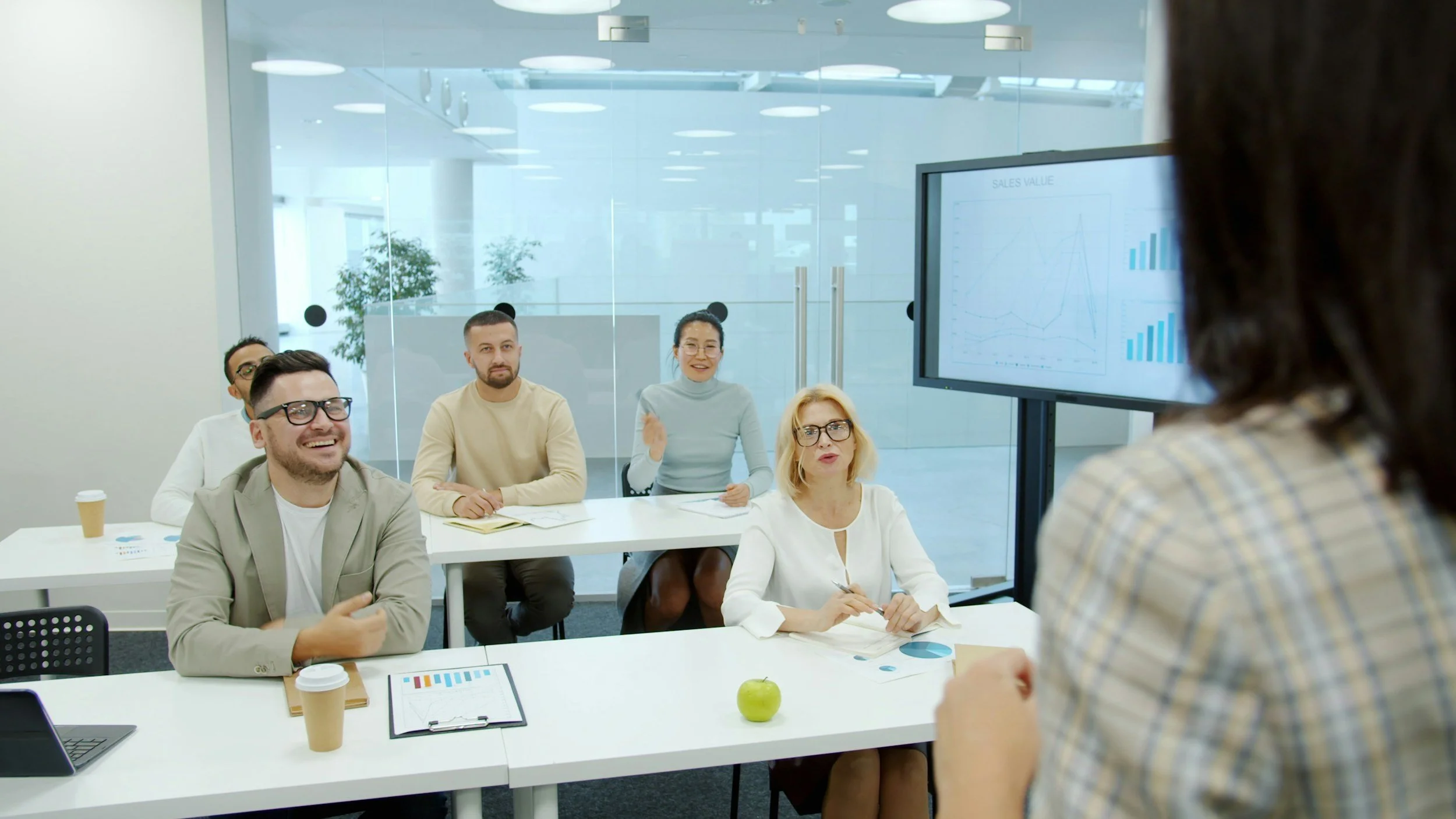 A business meeting with five people sitting at a white table with papers, drinks, and a green apple, listening to a woman speaking in front of a presentation screen displaying charts.