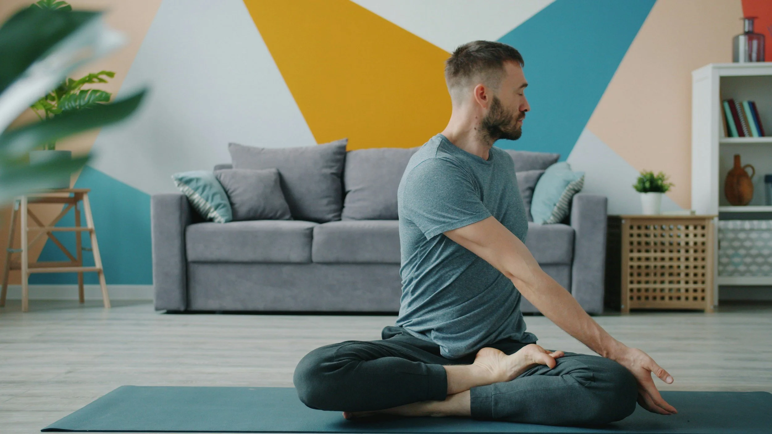 A man practicing yoga in a living room, seated in a cross-legged pose with his eyes closed and hands resting on his knees.