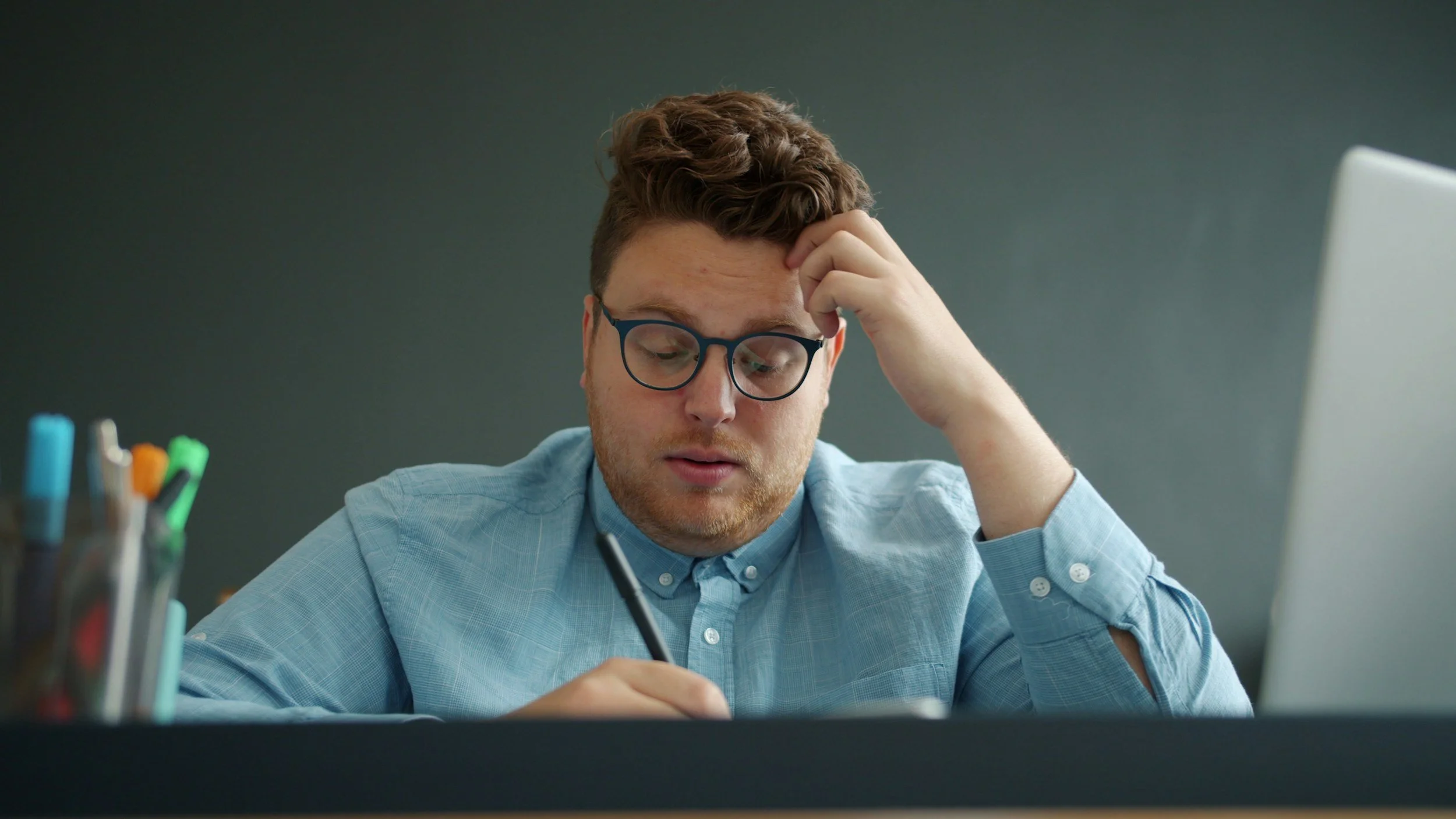 A young man with glasses, light blue shirt, and brown hair sitting at a desk, appearing stressed while holding a pen and writing, with a computer monitor in front of him and a container of colorful pens on the side.
