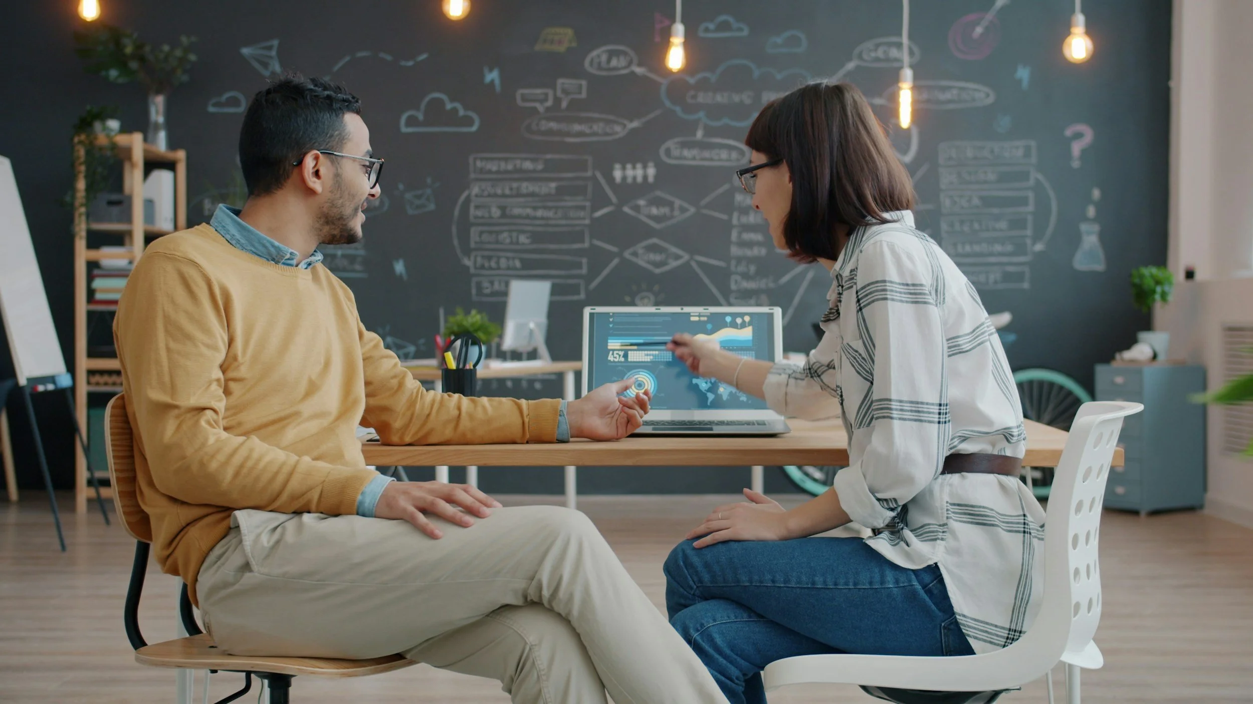 Two people, a man and a woman, are having a meeting in a modern office space. The man is wearing a yellow sweater and beige pants, and the woman is wearing a white plaid shirt and blue jeans. They are discussing with a laptop displaying graphs and data. The background features a black chalkboard wall with diagrams and doodles, pendant lights, and office furniture.