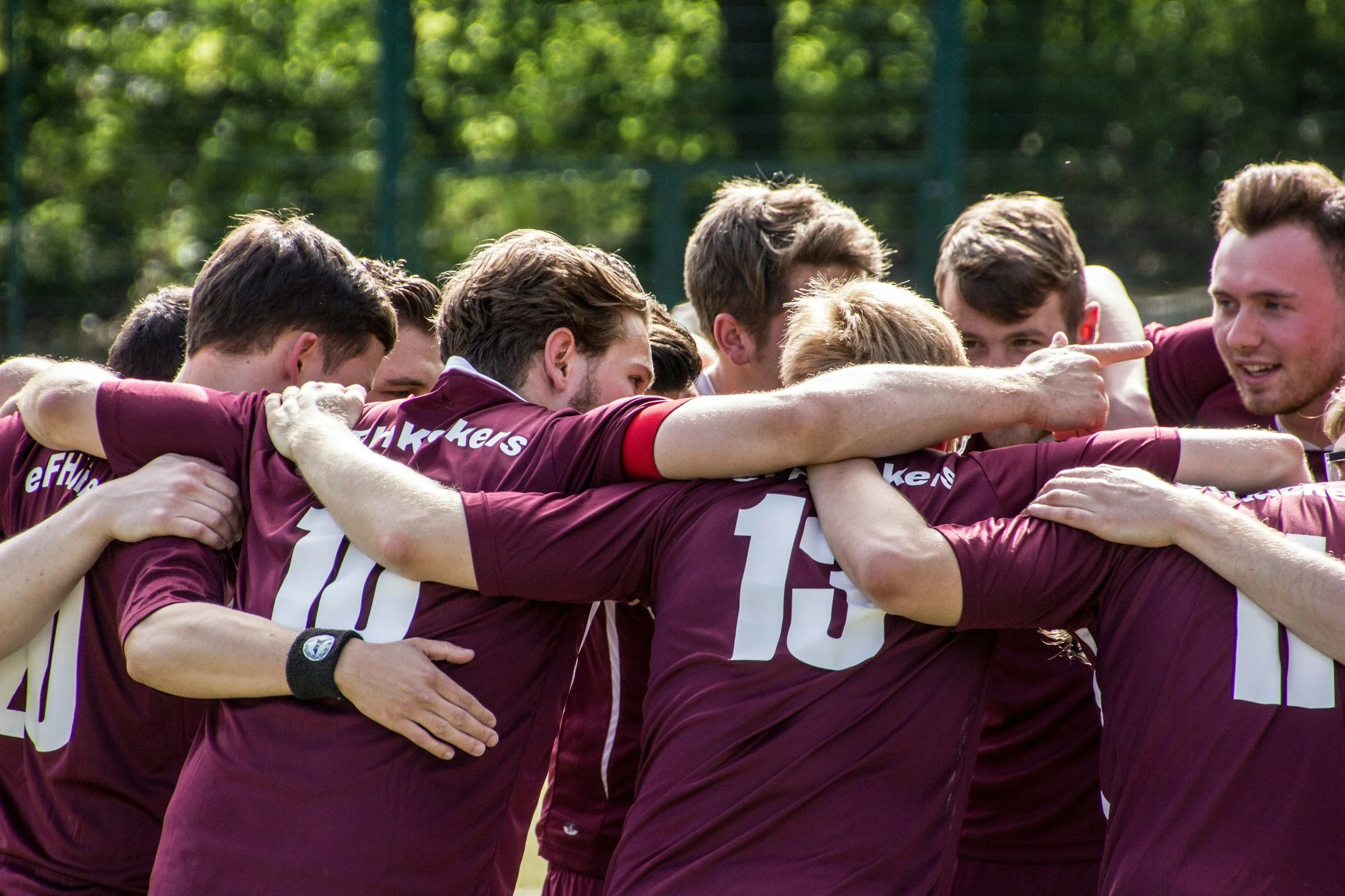 Group of male soccer players in maroon jerseys huddled together, arms around each other, during a match or practice.