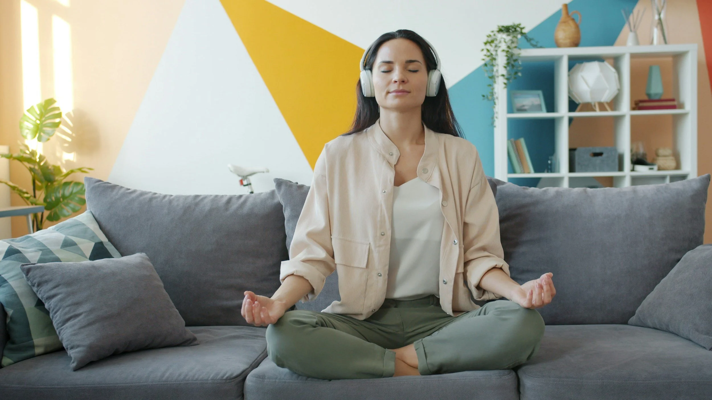 Woman sitting cross-legged on a gray couch in a meditative pose with eyes closed, wearing headphones in a colorful living room.