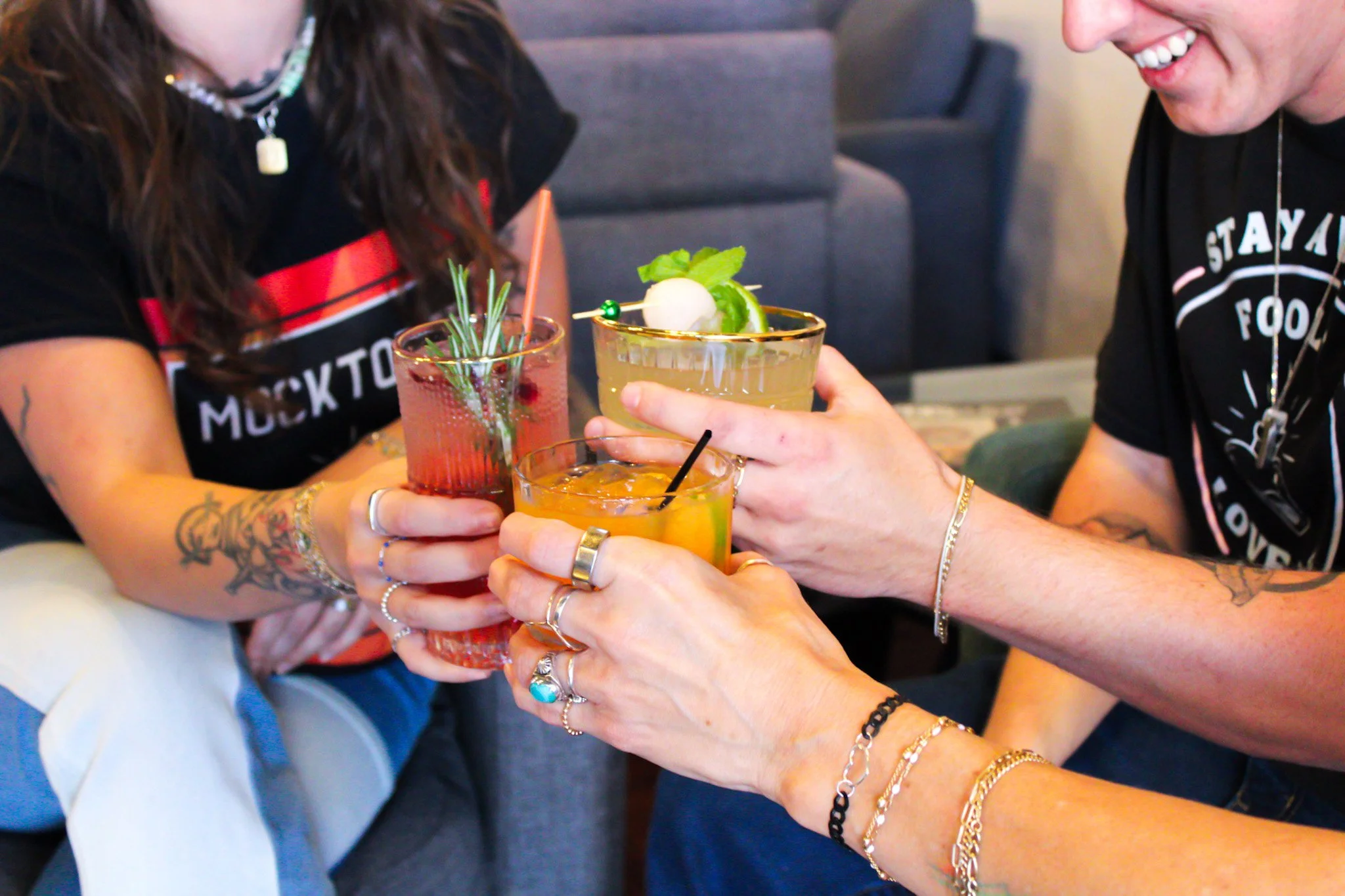 Two women holding colorful cocktails in a toast, one with a slice of lime and a cherry garnish, in a friendly social setting.