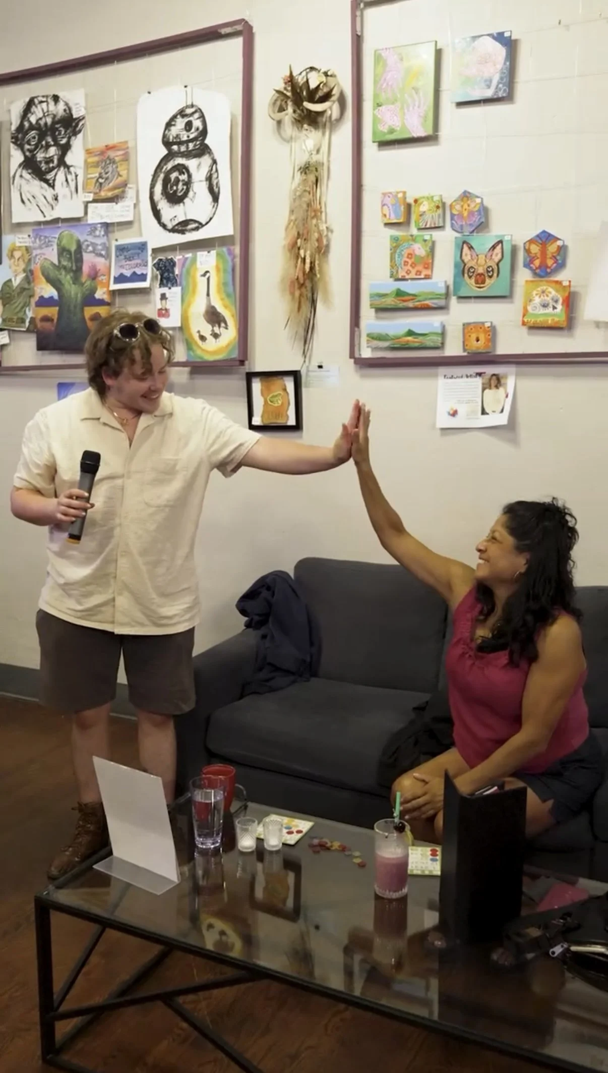 Two women in a living room high five during a social gathering. The woman on the left is holding a microphone and wearing sunglasses on her head, while the woman on the right is seated on a black sofa, smiling. The background features a wall with colorful artwork and a decorative hanging piece.