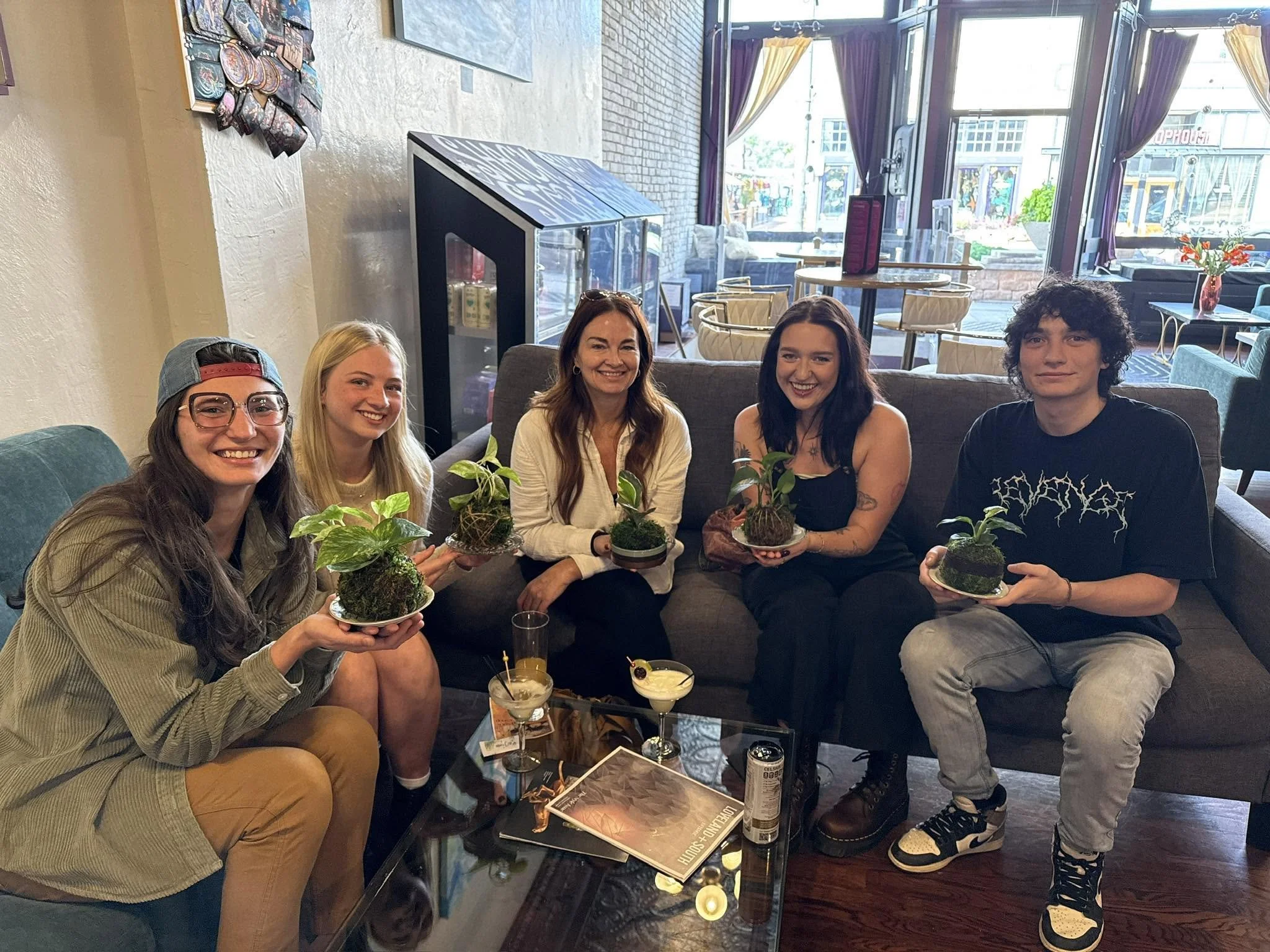 Five people smiling and sitting in a cozy cafe, holding small potted plants, with drinks on the table. Behind them are large windows with purple curtains, allowing natural light to fill the space.