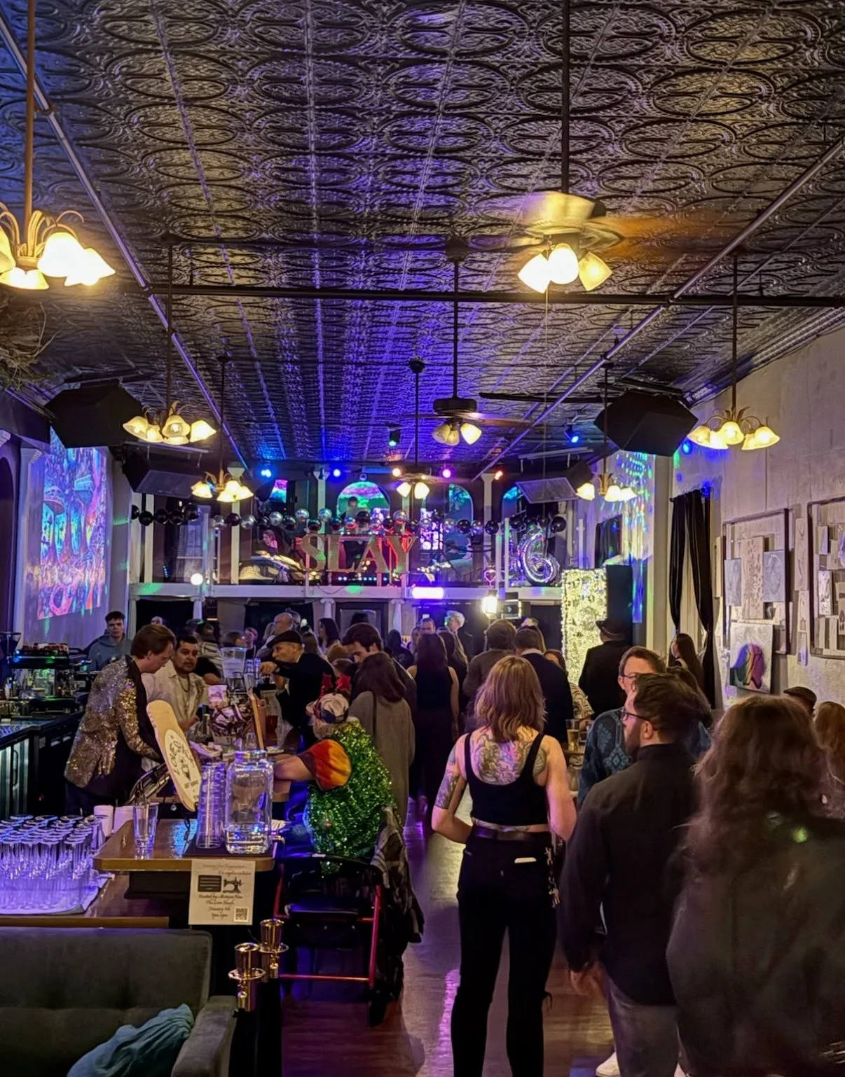 An indoor party scene with many people gathered, some wearing colorful and sparkly outfits, in a decorated venue with artwork on the walls, colorful lighting, and a sign that says 'SLAY' in the background.