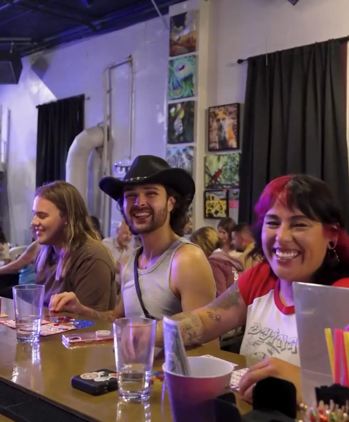 People sitting at a table in a social gathering or restaurant, smiling and laughing. One person is wearing a black cowboy hat, others are engaged in conversation. The background shows paintings on the wall and black curtains.
