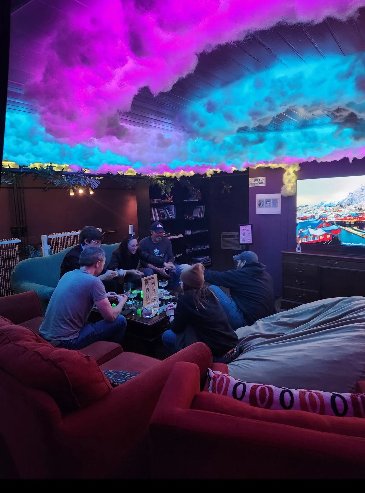 A group of people sitting on sofas and chairs in a cozy, dimly lit room with a colorful cloud-themed ceiling. They appear to be playing a tabletop game, with a TV screen displaying colorful scenery in the background.
