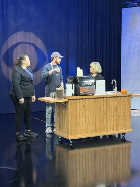Three people engaged in conversation behind a small wooden coffee cart on a stage with a blue curtain background.