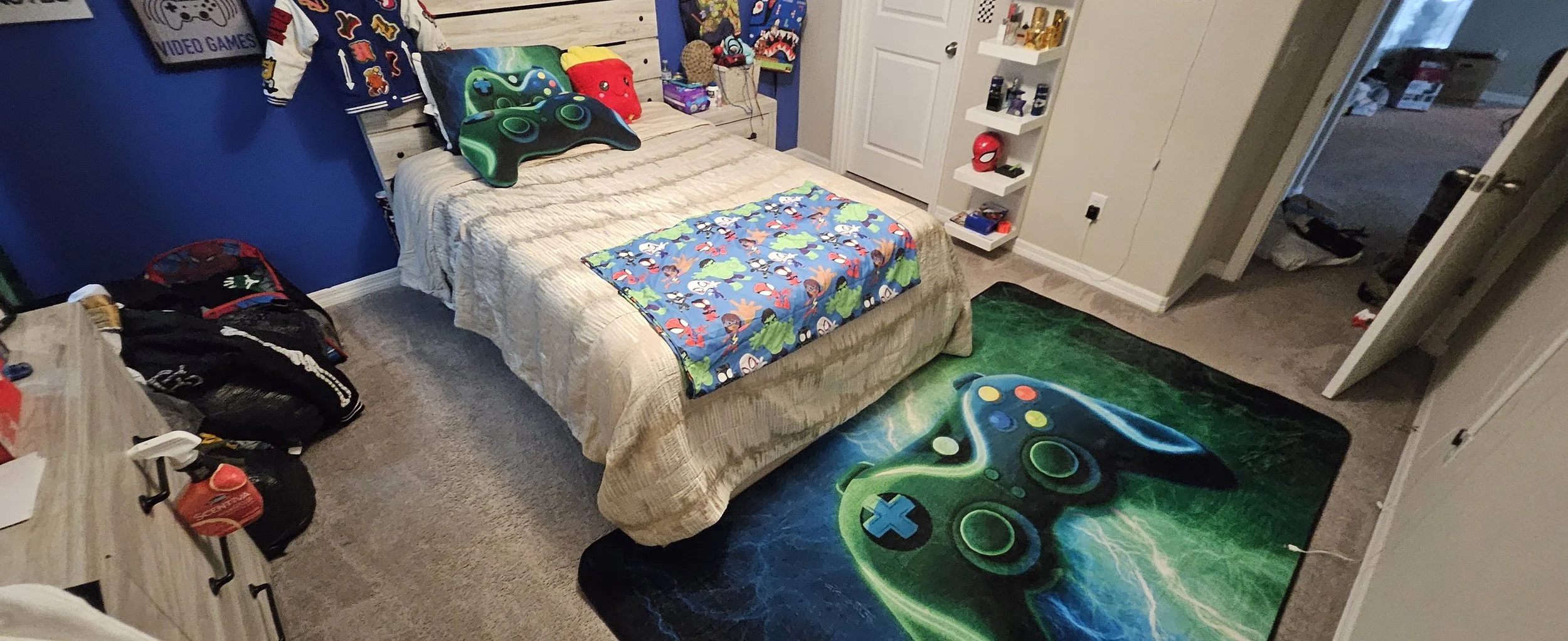Child's bedroom with a bed covered in a beige blanket, decorated with gaming-themed pillows, a colorful blanket at the foot of the bed, and gaming merchandise on a dresser. The room has a blue and beige wall, white shelving with collectibles, and a g