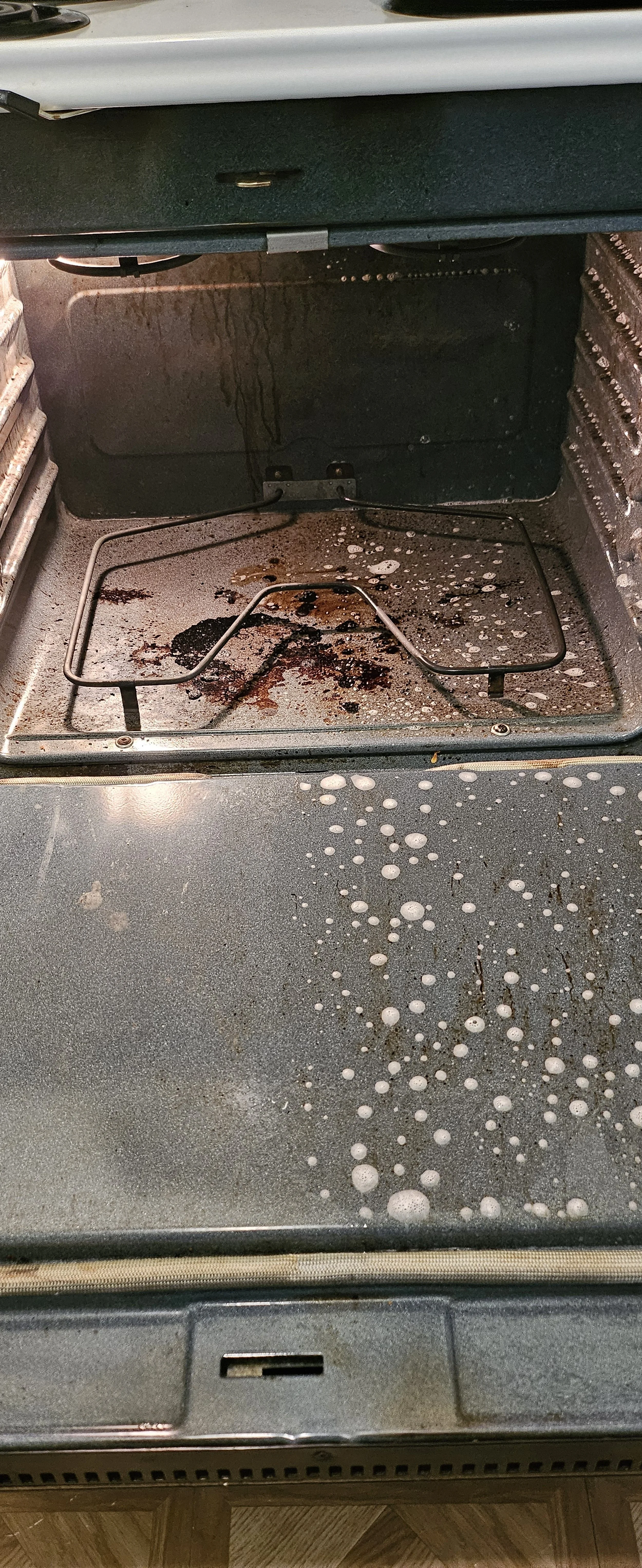 Inside an empty oven with burned and melted food residue on the oven floor and walls, and a metal rack inside.