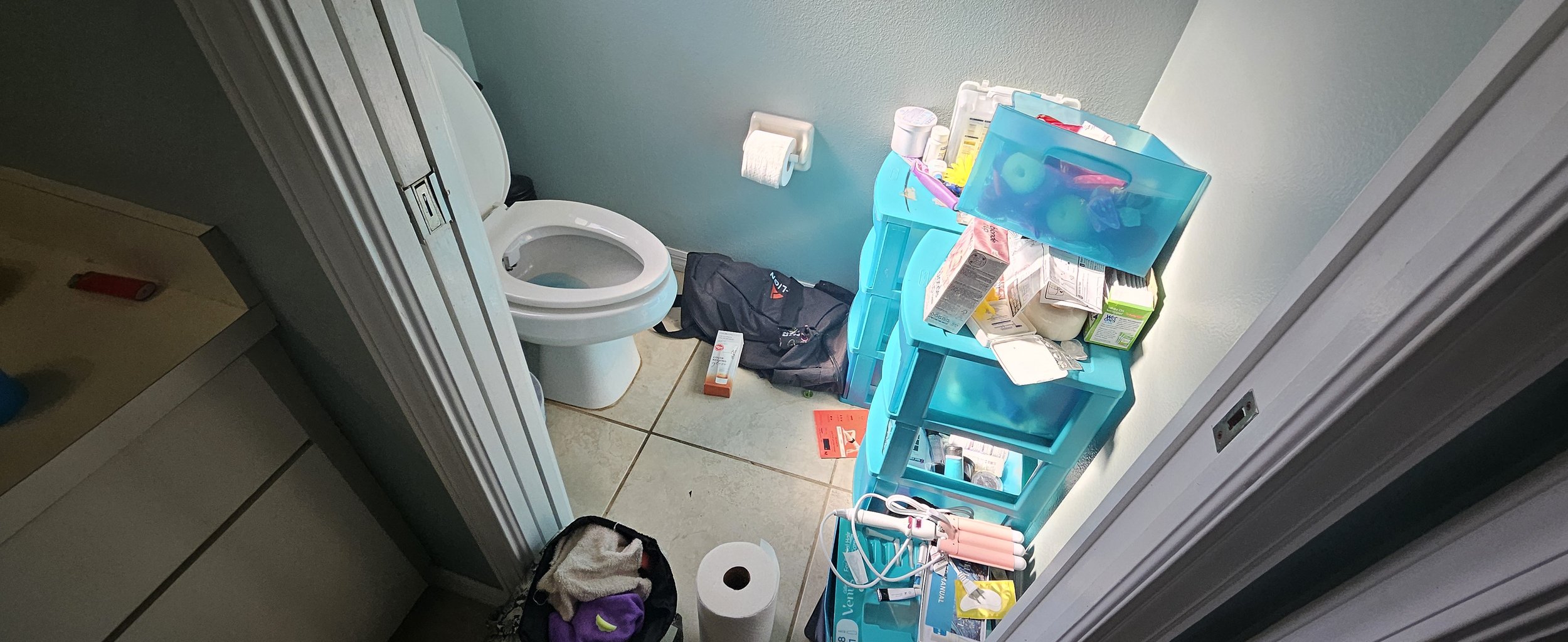 A cluttered bathroom corner with a toilet, several blue plastic storage drawers filled with medical supplies, a black backpack on the floor, a roll of toilet paper, and various medical items and packaging on top of the drawers.