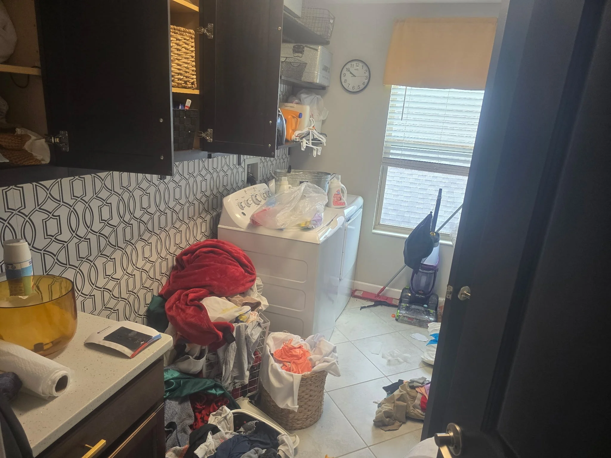 Messy laundry room with clothes scattered on the floor and laundry basket, washer, and cabinets visible.