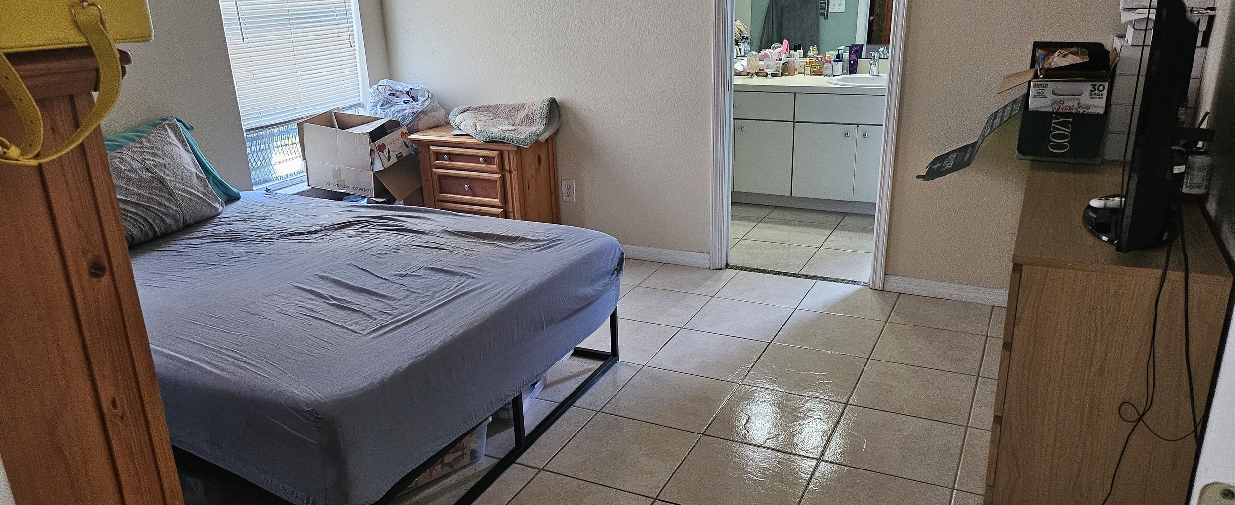 A modest bedroom with beige tiled floor, a bed with gray bedding near a window with blinds, a wooden nightstand with a towel and some boxes, and a wooden TV stand with a small TV and assorted items. An open door reveals a bathroom with white cabinetr