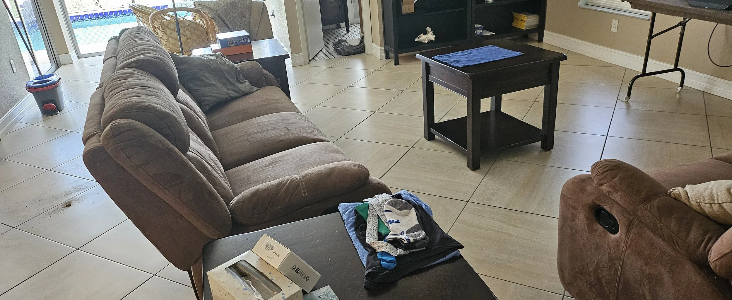 Living room with beige tiled floor, brown sofa with pillows, small black coffee table with clothes and a box on it, and sliding glass door leading outside. In the background, there is a black shelf, a blue towel on the coffee table, a brown recliner,