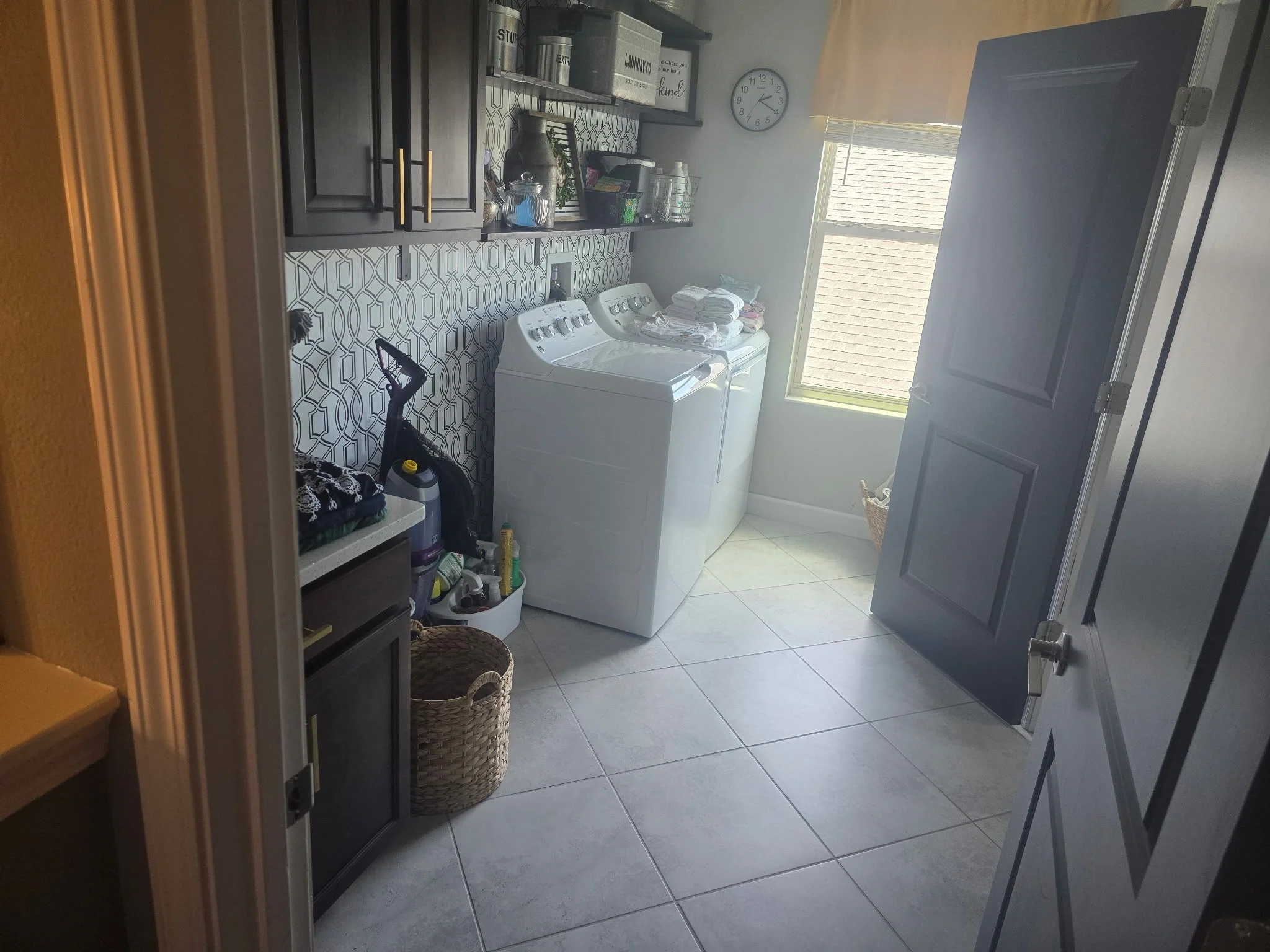 Laundry room with a washing machine and dryer, dark cabinets, shelves with laundry supplies, a window with sunlight, a clock, baskets, and cleaning tools.
