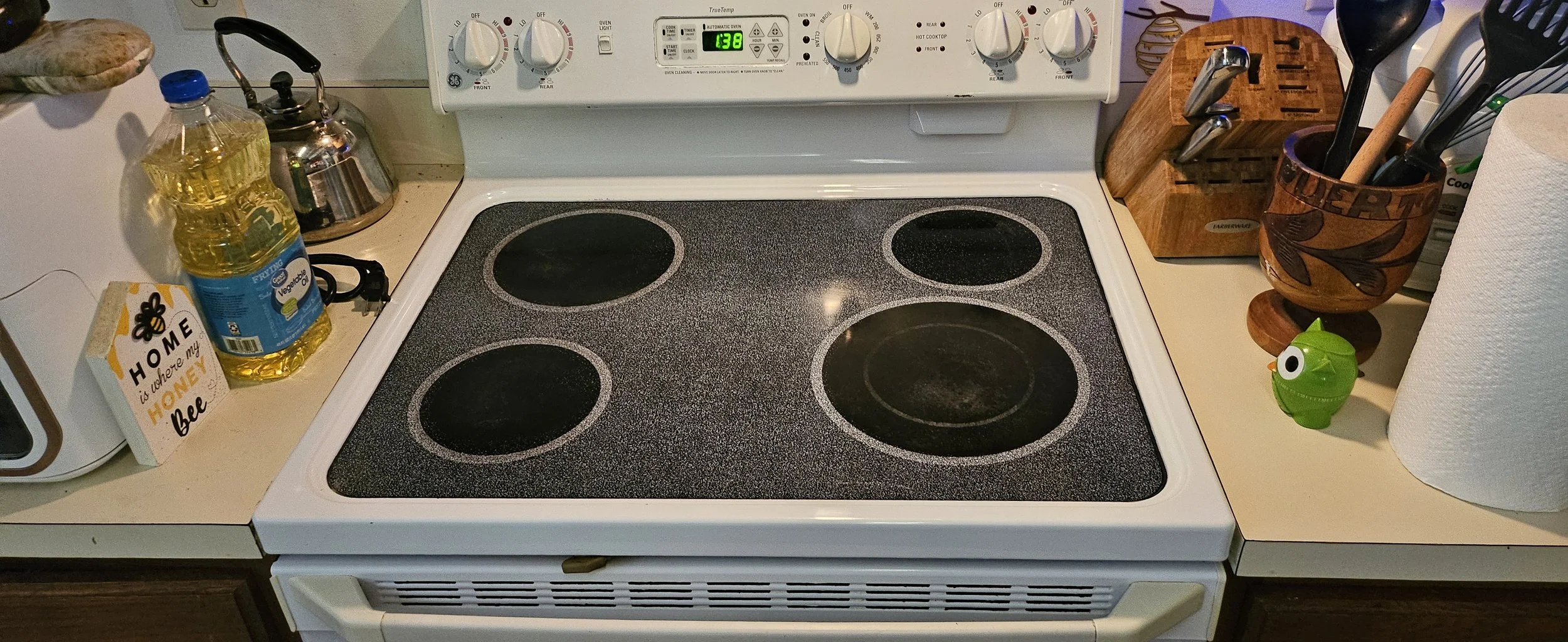 White electric stovetop with four black burners, surrounded by kitchen items including a bottle of oil, a tea kettle, a wooden knife block, a green toy, and paper towels.