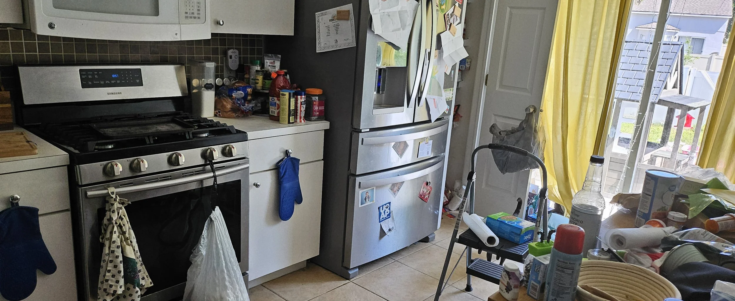 A cluttered kitchen with a stainless steel refrigerator, a stove, and a countertop filled with various food items and mess. There is a yellow curtain over a sliding glass door leading outside, and a small step stool with papers and a roll of paper to
