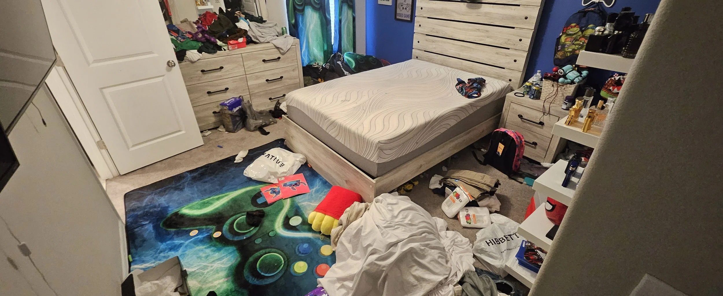 A cluttered bedroom with an unmade bed, piles of clothes, scattered items, and various household objects on the floor and around the room.