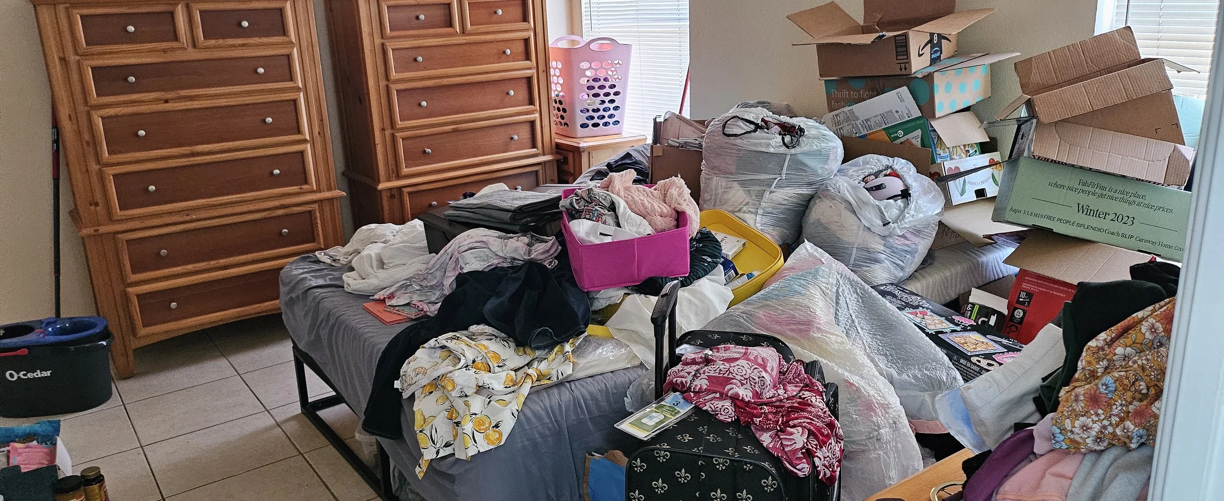 Messy bedroom with a bed covered in clothes, boxes, and various household items, wooden dressers along the wall, and cluttered surfaces.